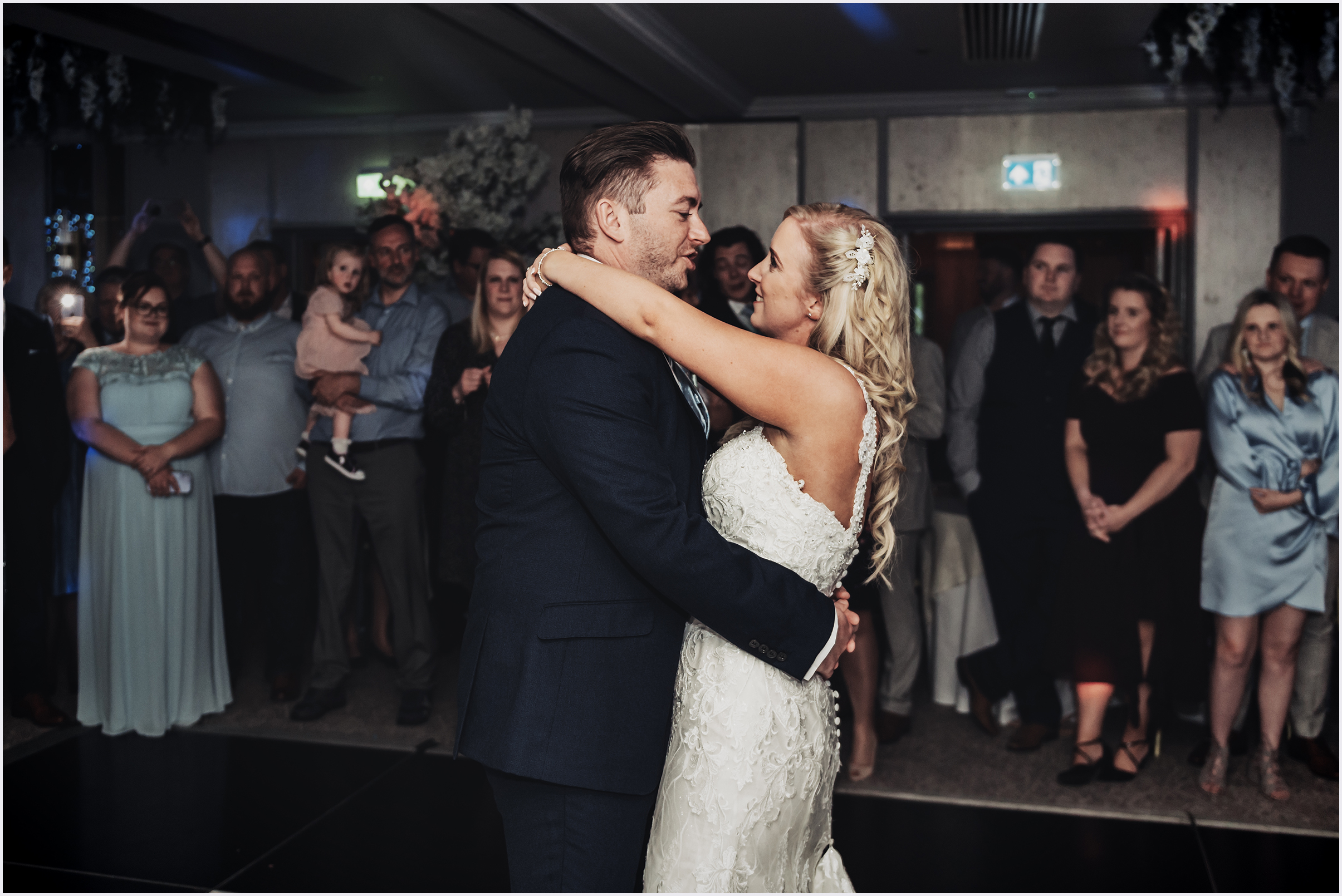 A bride drapes her arms around her new husband's shoulders as he has his arms around her waist as guests watch them dance their first dance at The Grosvenor Pulford Hotel and Spa.