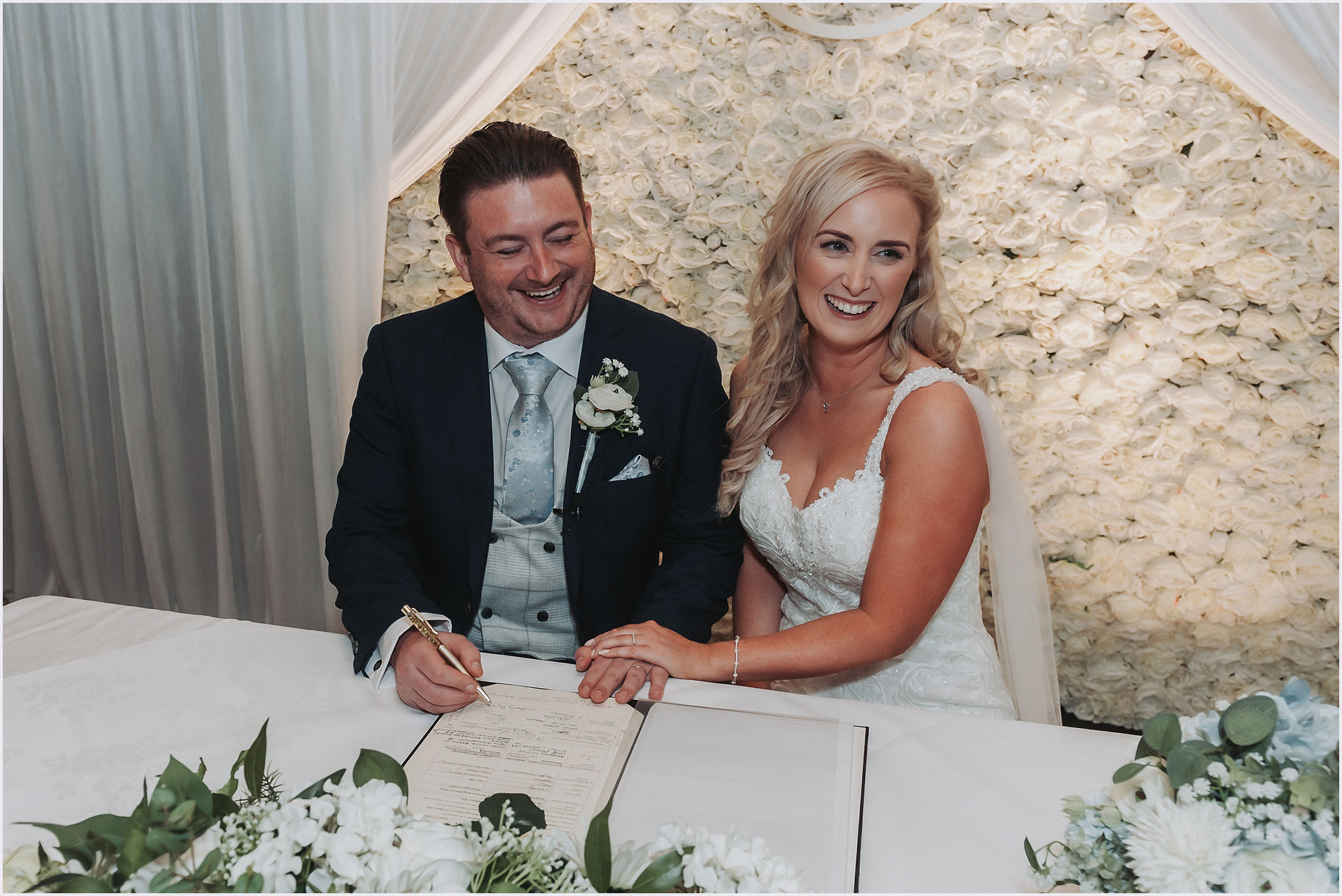 A bride gently rests her hand on her new husbands hand after signing the register at their wedding.  Both are laughing.