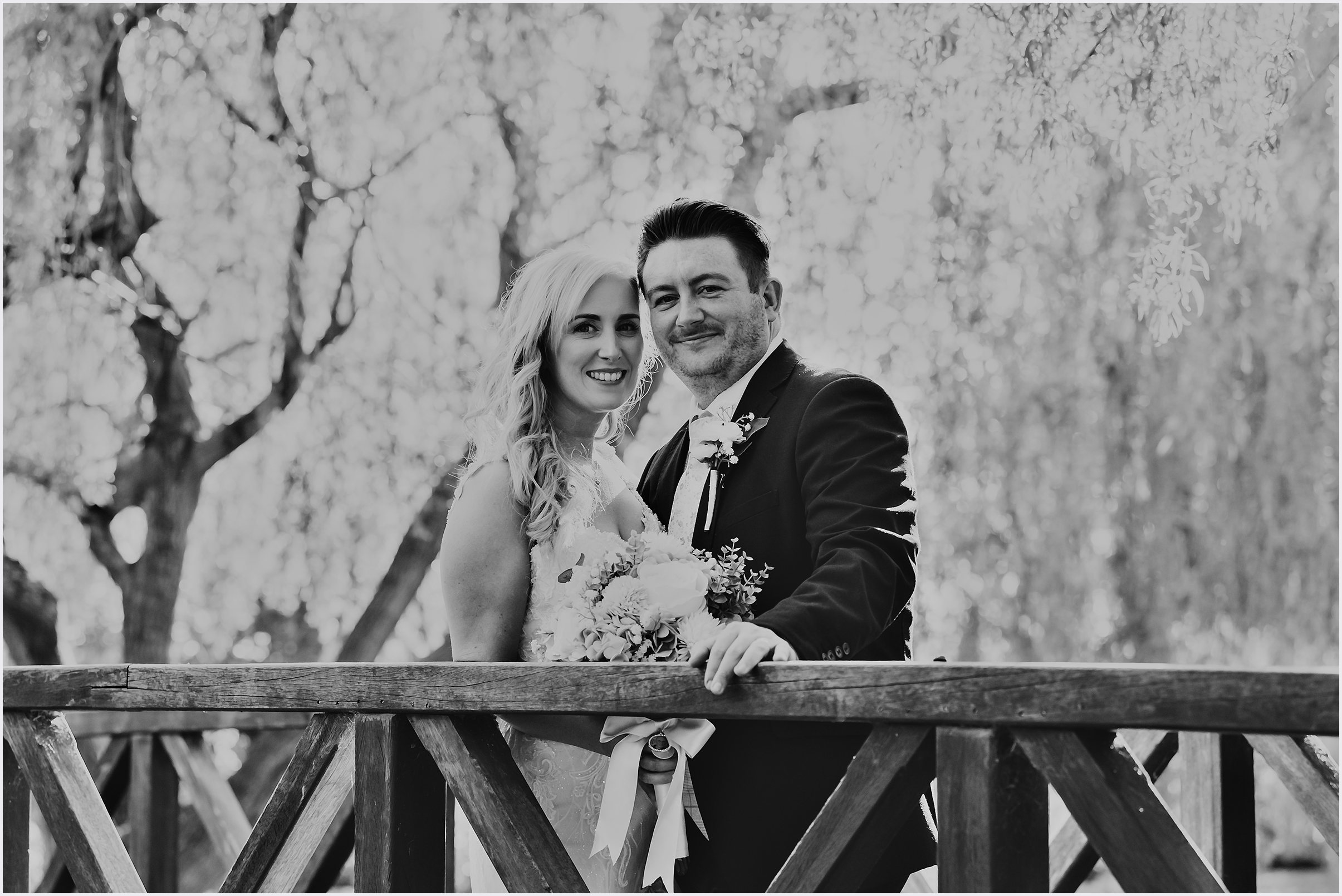 A groom's hand rests on the railing of a wooden bridge as he stands next to his new wife smiling at the camera during the bride and groom portraits at The Grosvenor Pulford Hotel and Spa.