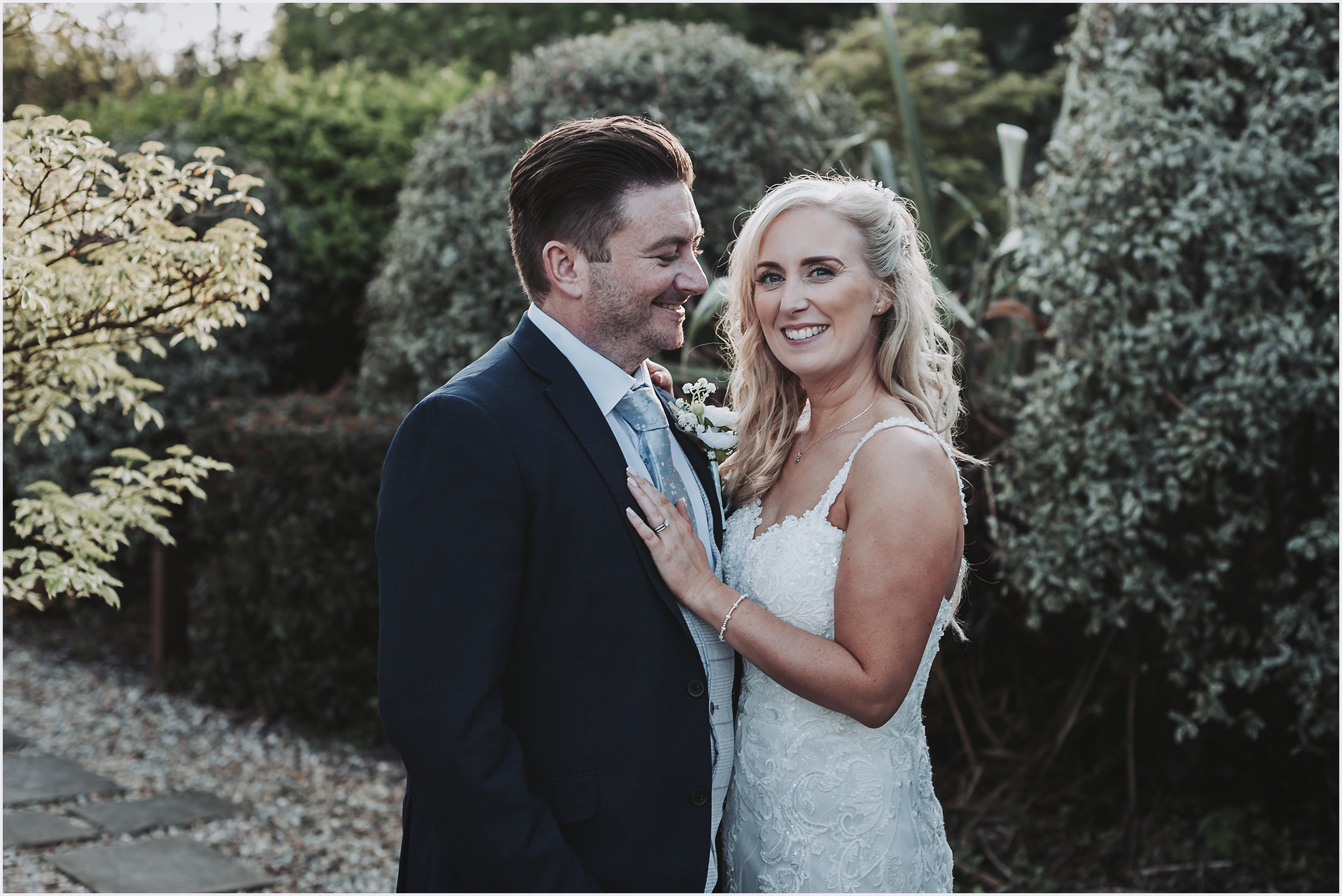 A bride resting her hand on her new husband's shoulder as she smiles at the camera in the beautiful gardens at The Grosvenor Pulford Hotel and Spa.