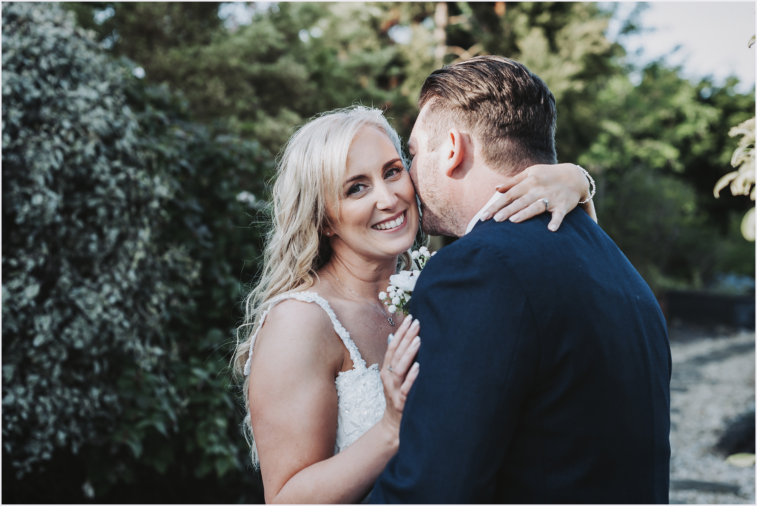 A groom whispers into his wife's ear as she smiles at the camera in the gorgeous gardens at The Grosvenor Pulford Hotel and Spa.