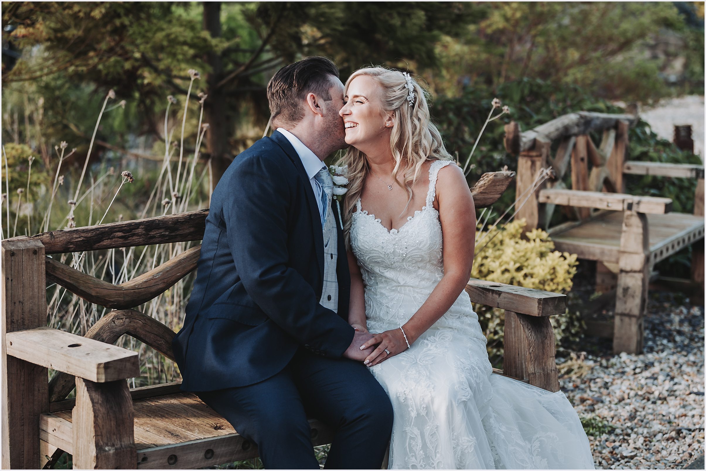 A groom is whispering into his new wife's ear making her laugh while sitting on a rustic wooden bench in the Japanese Gardens at The Grosvenor Pulford Hotel and Spa