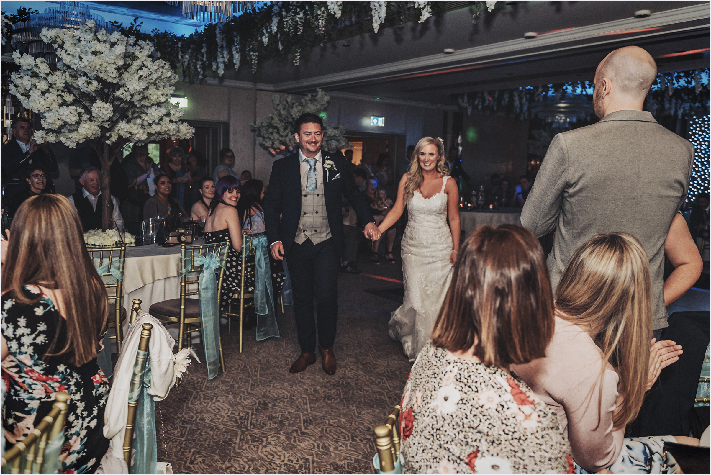 A bride and groom walk happily hand in hand towards the dance floor for their first dance.  Guests smiling at them.  