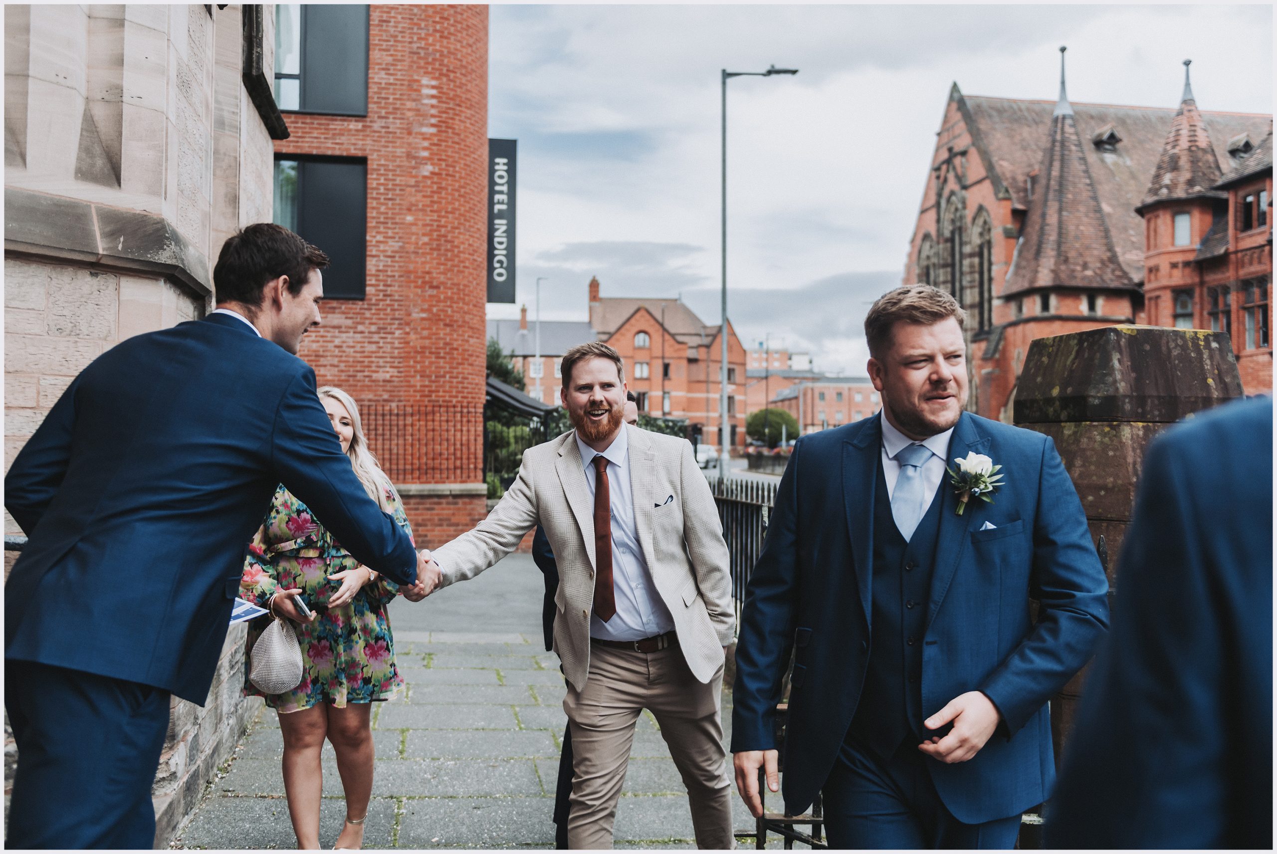 Guests shake hands and greet each other outside a church before their friends get married in Chester City Centre.  Image captured by Helena Jayne Photography Chester Wedding Photographer