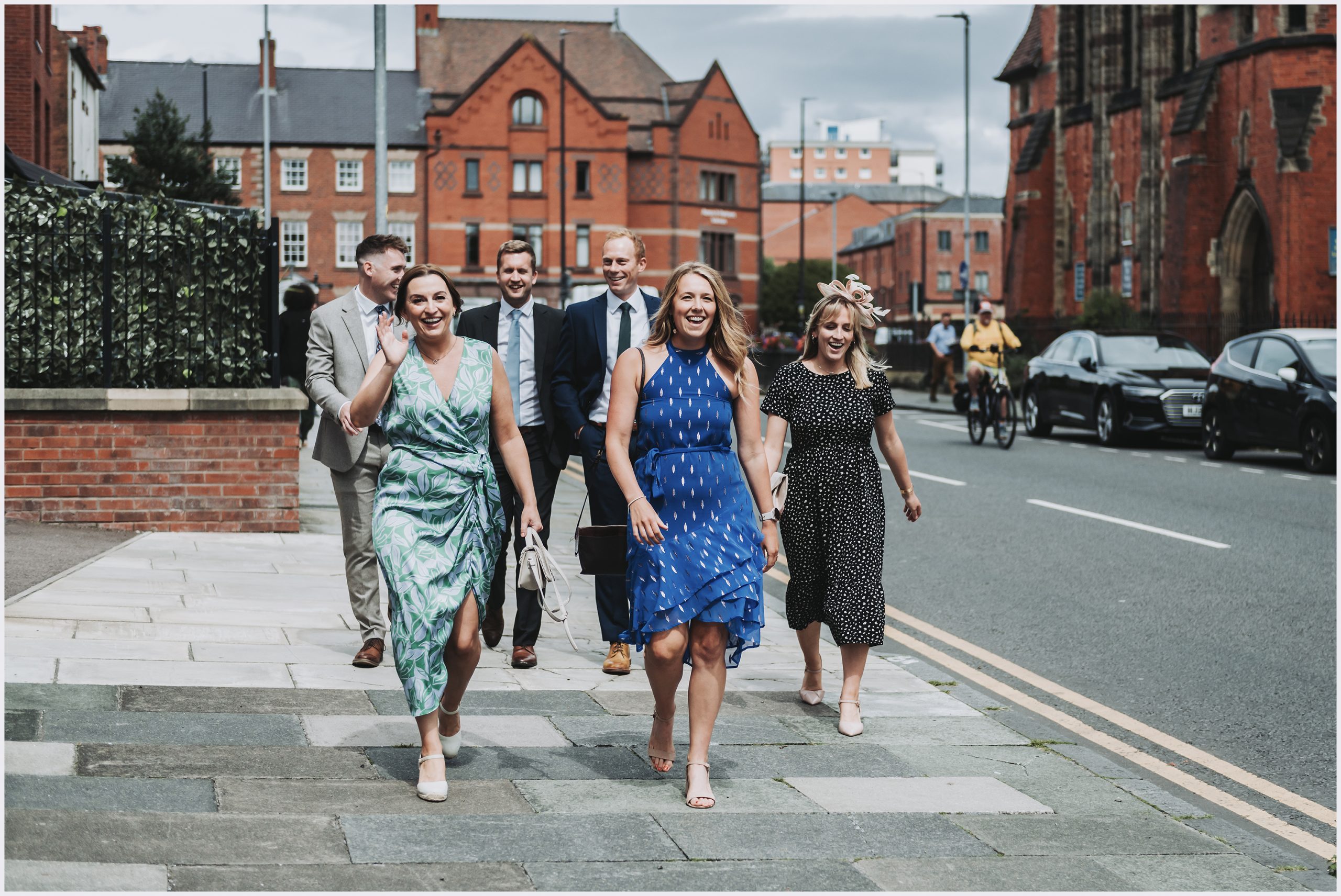 A group of friends dressed in beautiful dresses walk towards the church waving at their friends ahead of their wedding in Chester City Centre.  Image captured by North Wales wedding photographer Helena Jayne Photography