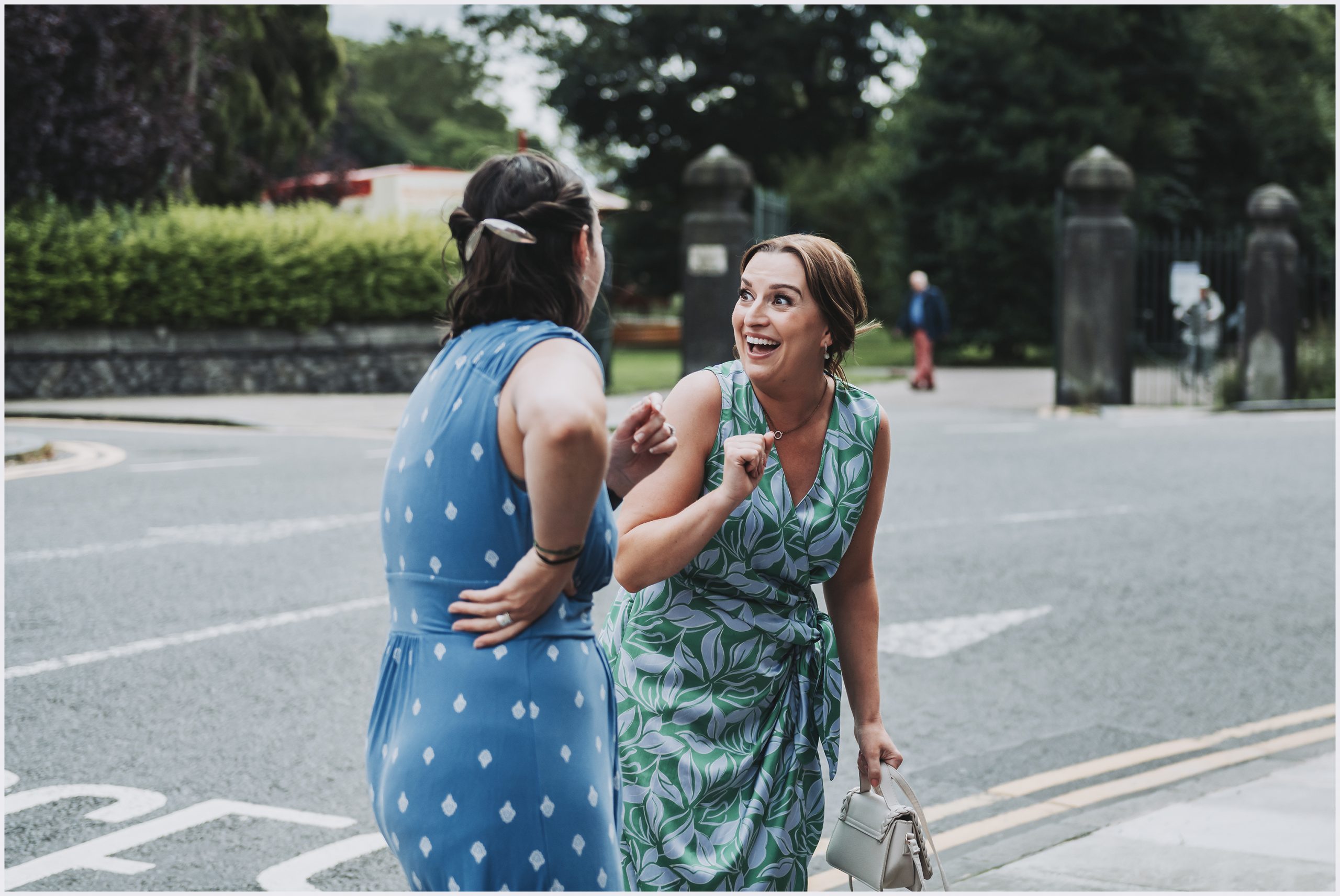 Two girl friends laugh and joke outside the church where their friends are about to get married.  Image captured by Helena Jayne Photography North Wales wedding photographer