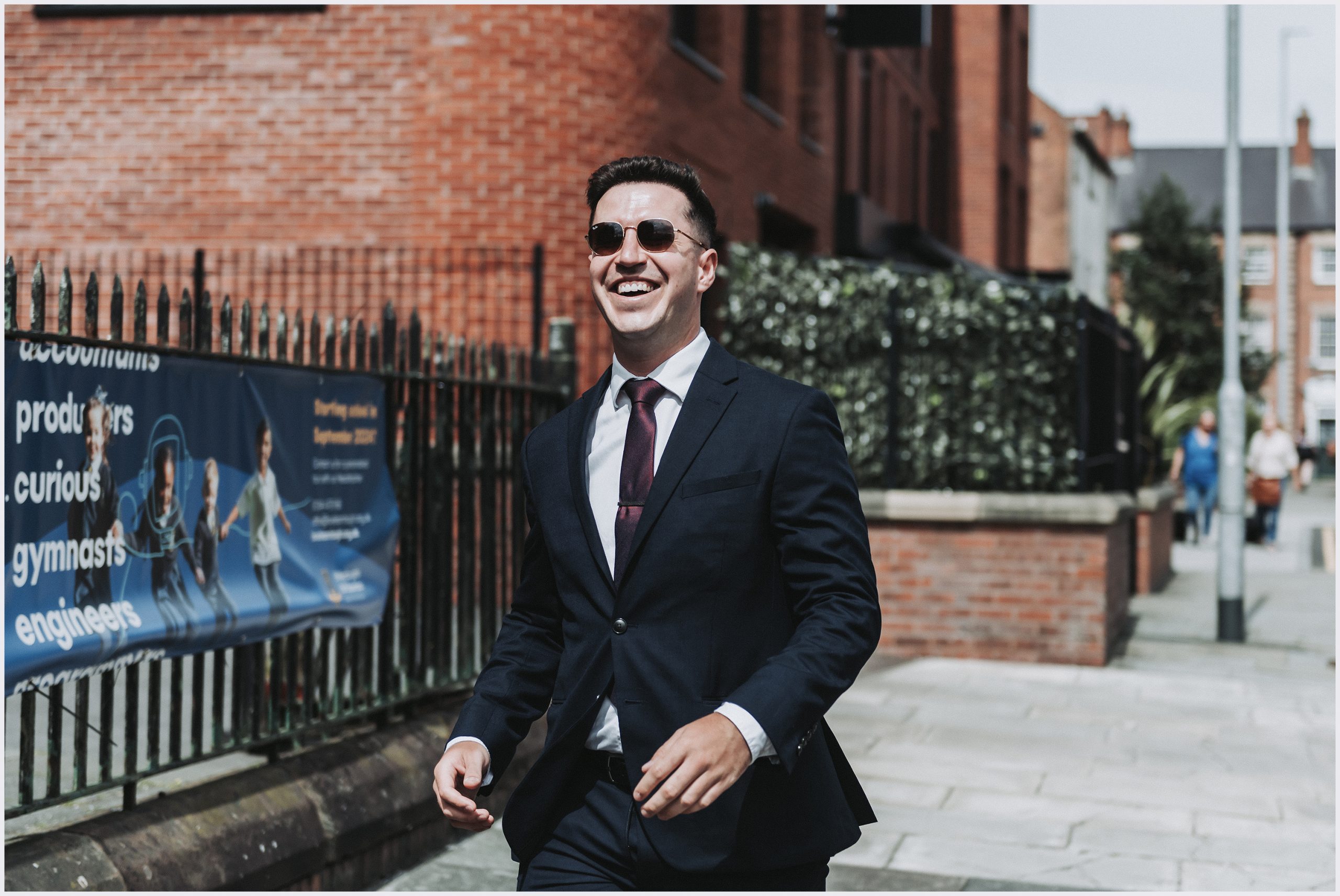 A smiling man wearing sunglasses and a really lovely dark suit smiles as we walks towards his friends who are waiting outside a church ahead of their friends wedding in Chester City Centre.  Image captured by Chester wedding photographer Helena Jayne Photography