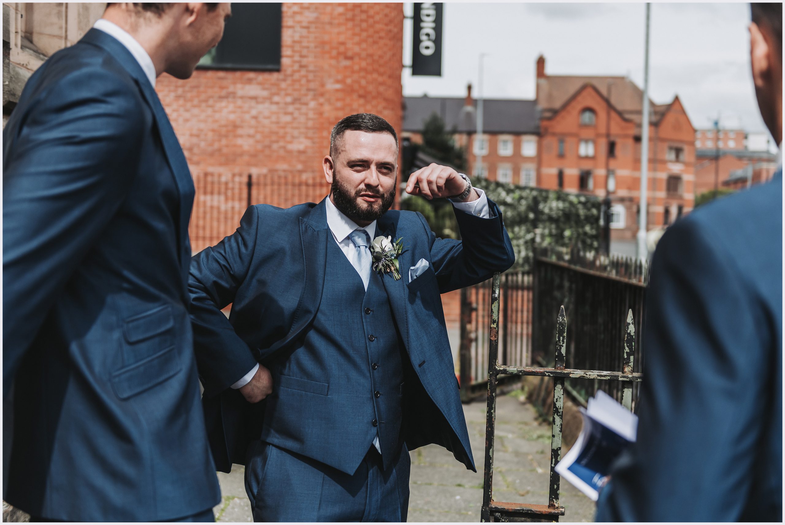 An usher wearing a blue suit casually leans his elbow against the church gate while greeting wedding guests into the church.  Image captured by north Wales wedding Photographer Helena Jayne Photography
