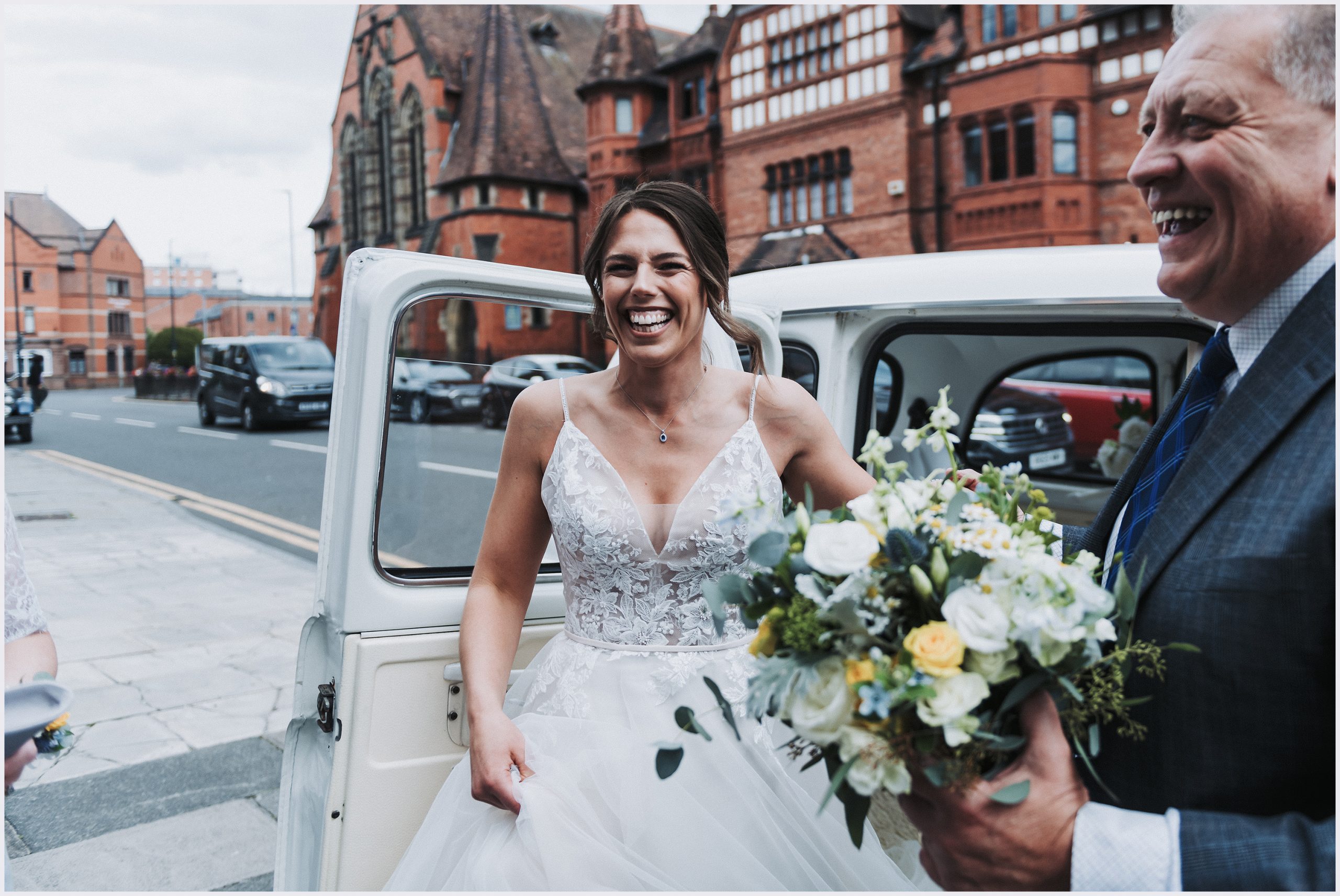 A beautiful smiling bride wearing her wedding gown steps out of the car that has brought her to the church to be married.  The driver helps her out carrying her bouquet.  Image captured by Helena Jayne Photography Chester wedding photographer