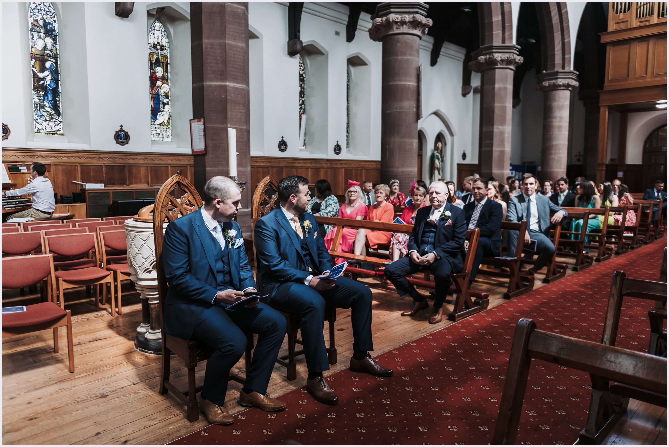 A groom and his best man sit in grand chairs at the top of the aisle of a church while the wedding guests watch on before the wedding ceremony