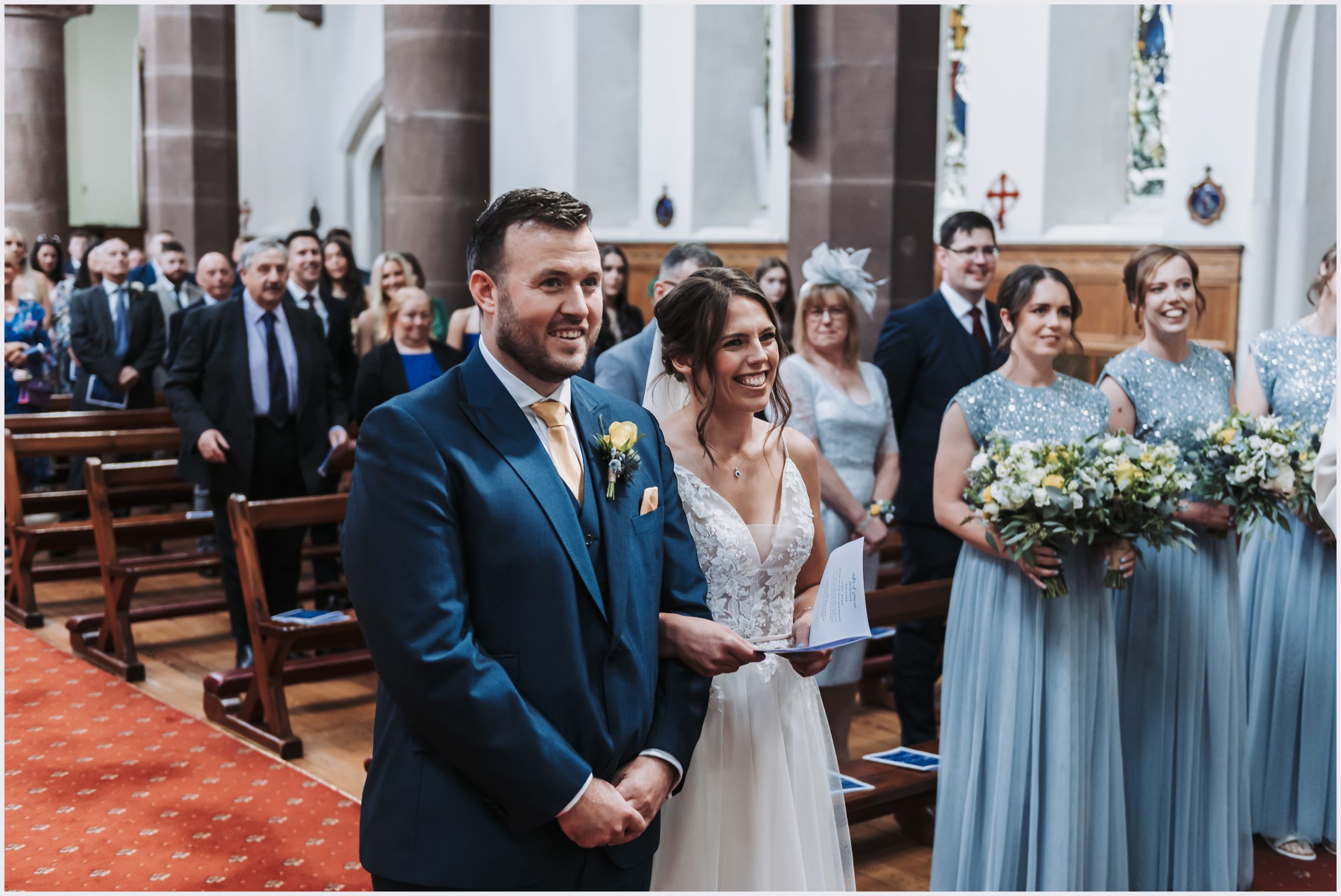 A beautiful bride and gorgeous groom stand side by side at the top of the aisle in a church during their wedding ceremony in Chester.