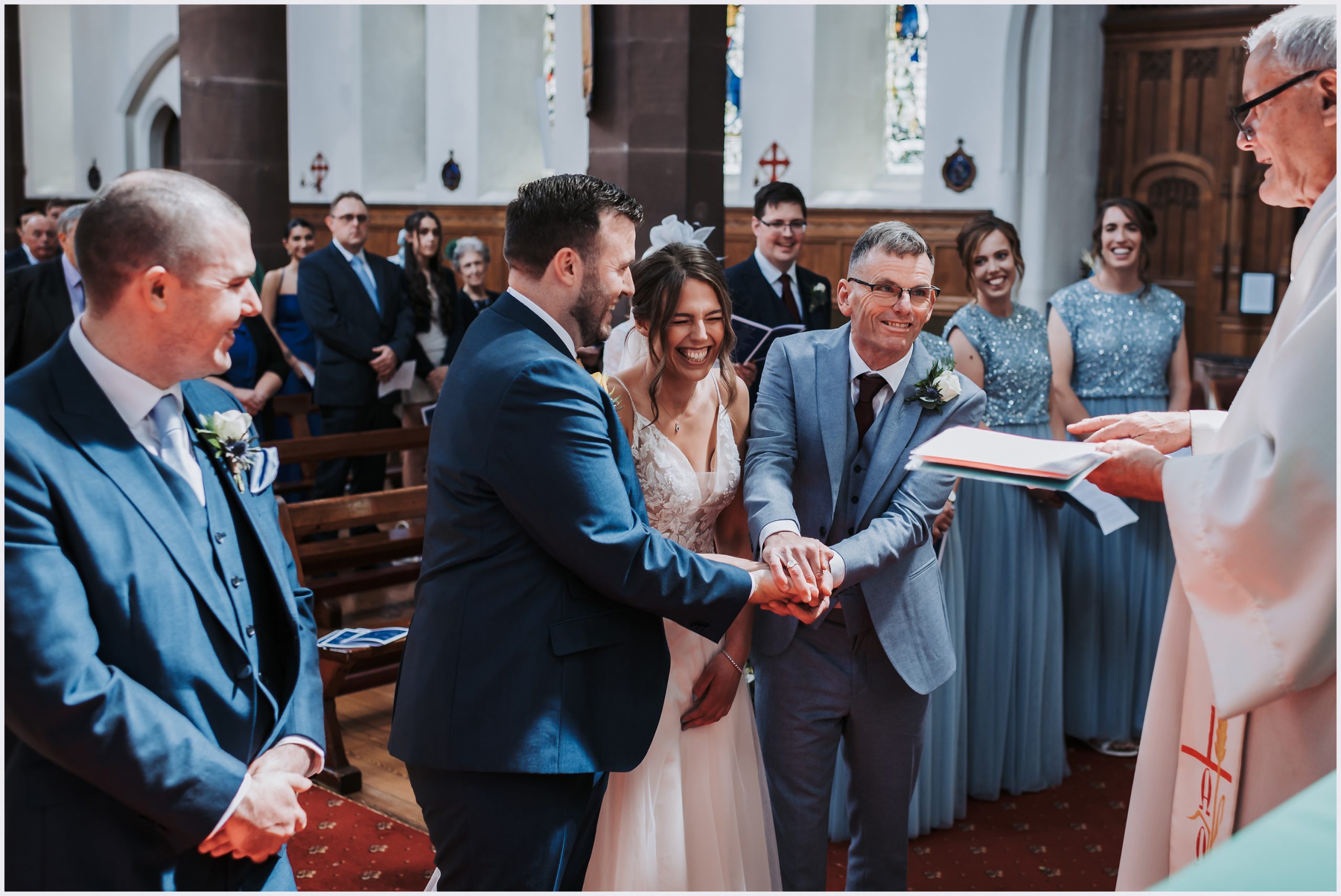 A laughing bride's hand is placed in to her husband's hand by her father during their wedding ceremony in Chester.  Image captured by Helena Jayne Photography a north Wales wedding photographer