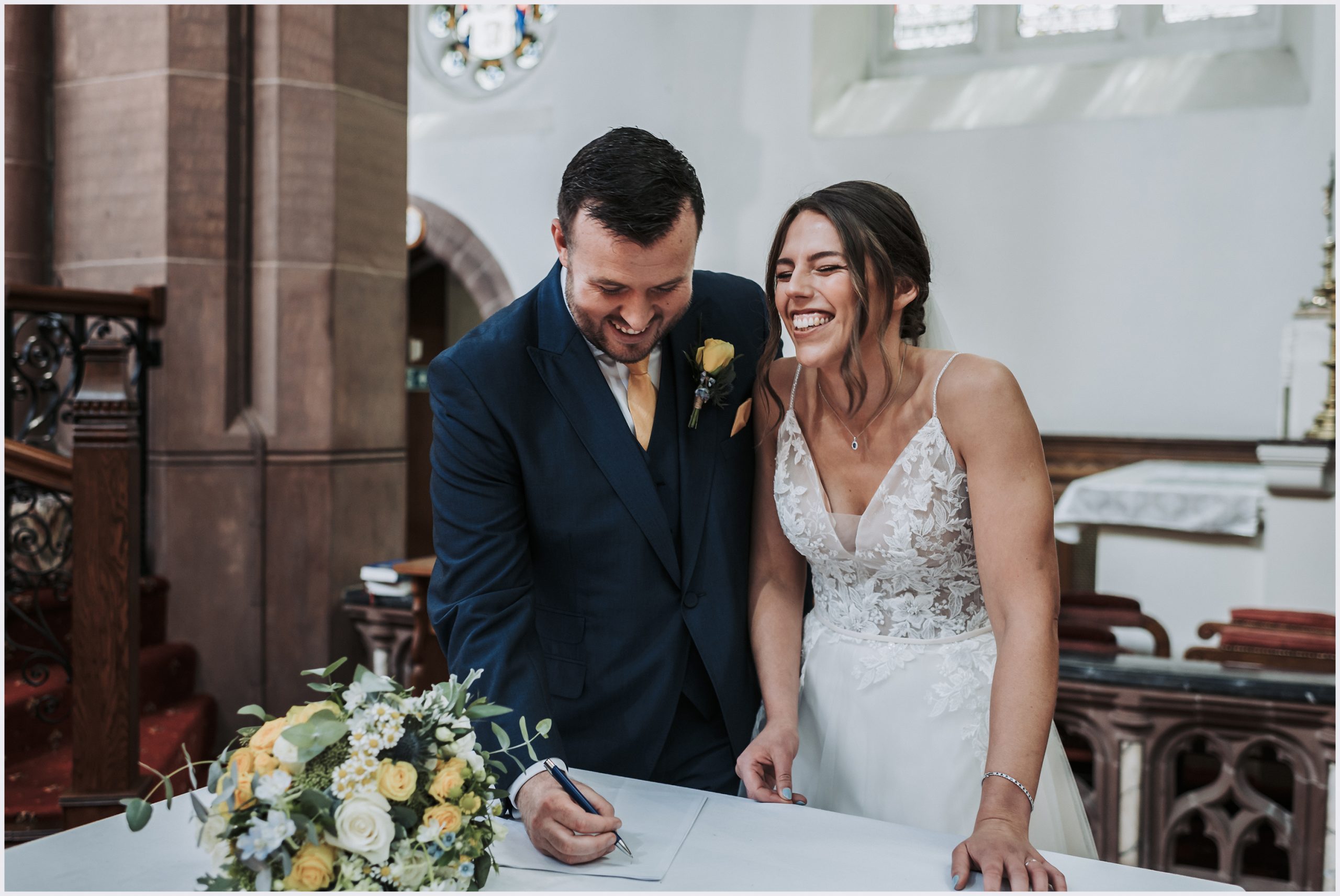 A bride and groom giggle as they sign the register after their church wedding ceremony.  Image captured by North Wales wedding photographer Helena Jayne Photography