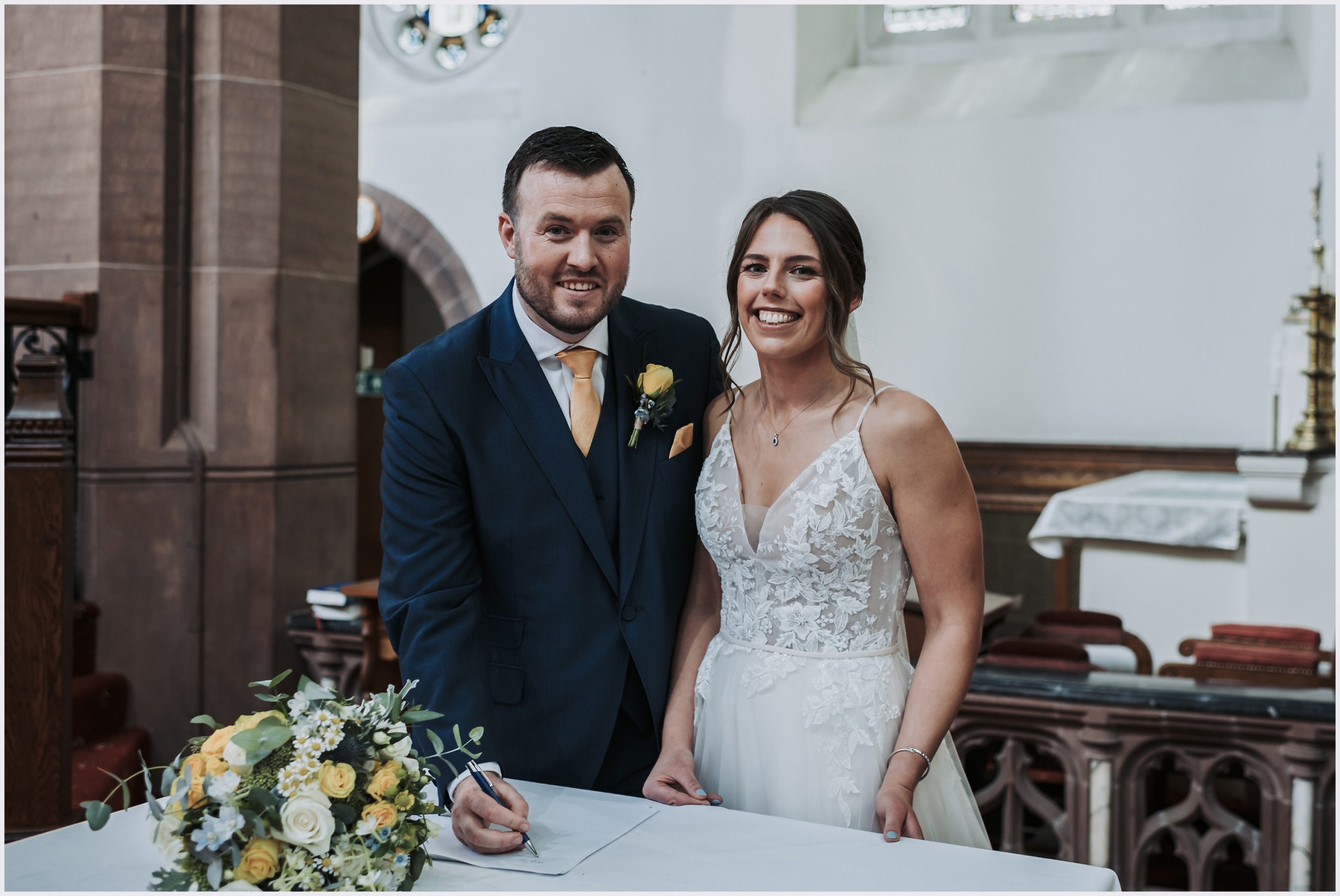 A bride and groom pose smiling at the camera after signing the register at their church wedding ceremony.  Image taken by Chester wedding photographer Helena Jayne Photography