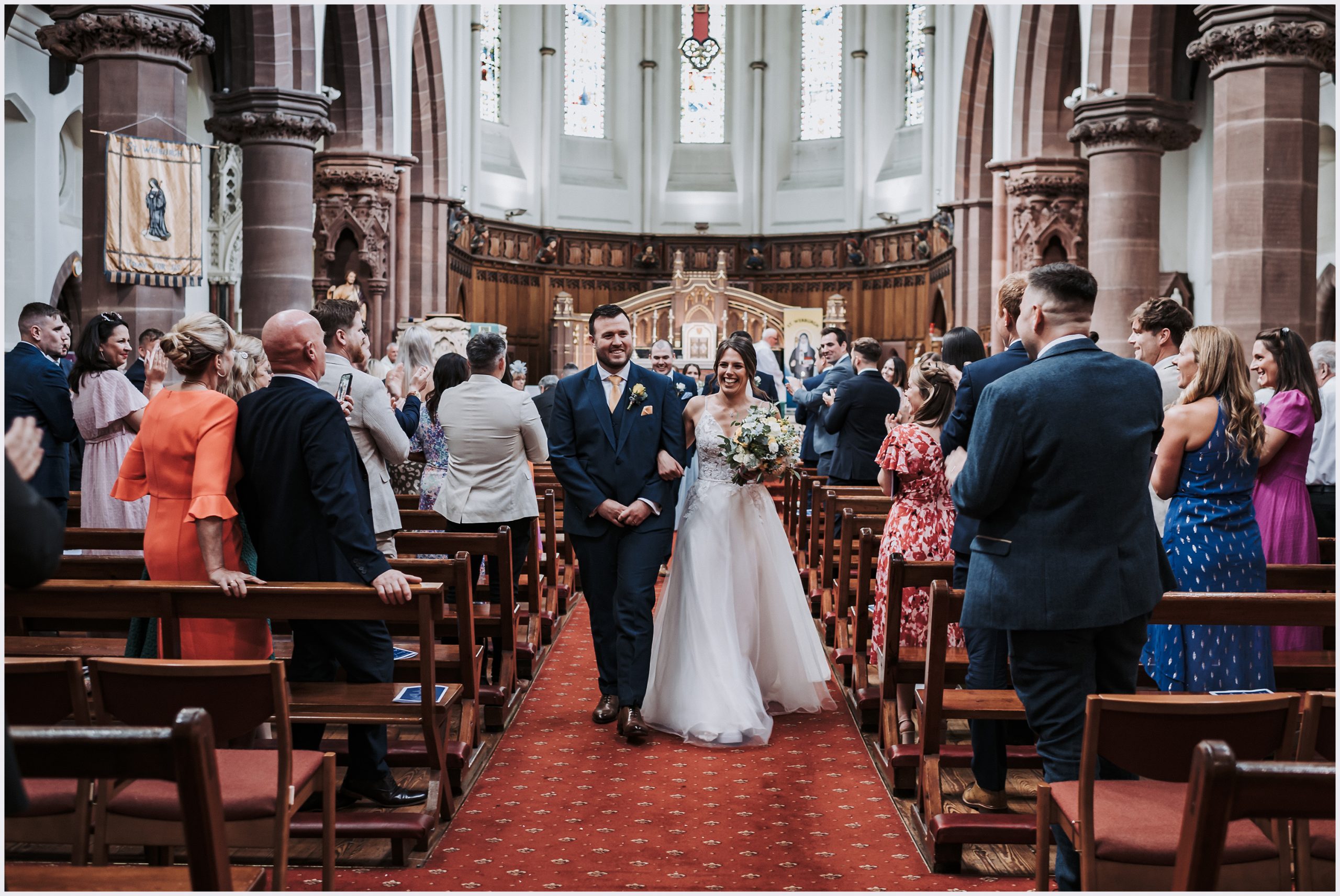 A wide shot of the back of the church as a the newly married smiling bride and groom walk outside after getting married.  Image captured by Helena Jayne Photography.