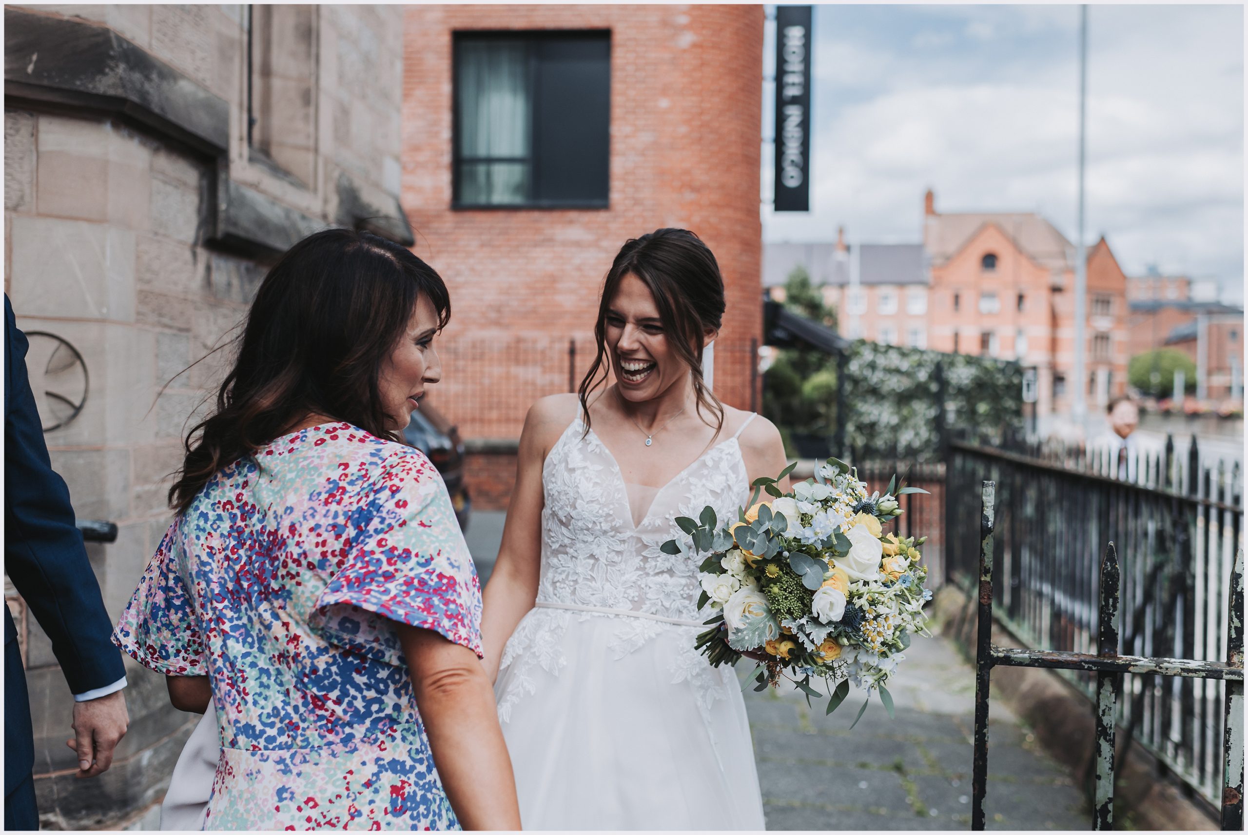 A bride and a friend share a joke smiling outside the church where she has just got married in Chester.  Image captured by Helena Jayne Photography a wedding photographer based in north Wales