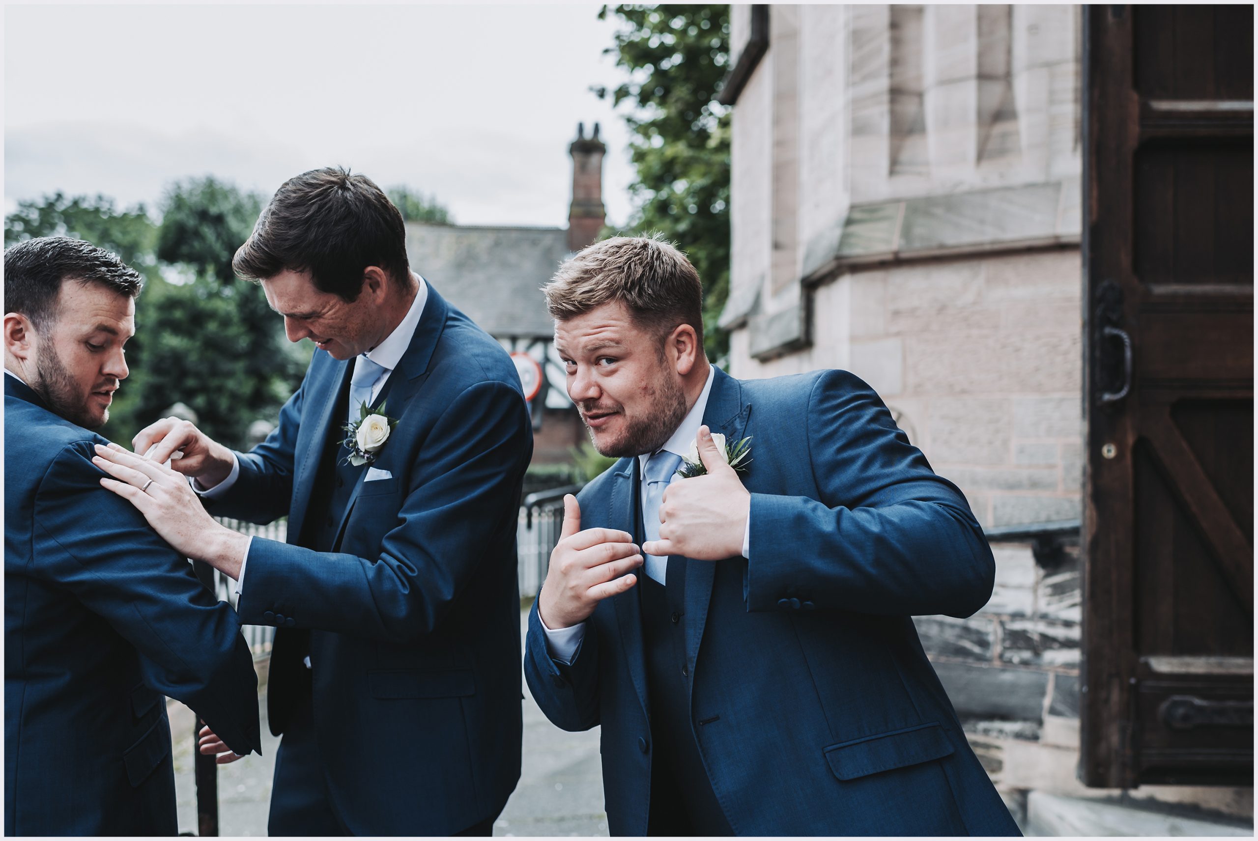 An usher rubs make up off the groom's jacket while another usher gives the thumbs up to the camera after a wedding ceremony in Chester.  Image captured by Chester wedding photographer Helena Jayne Photography