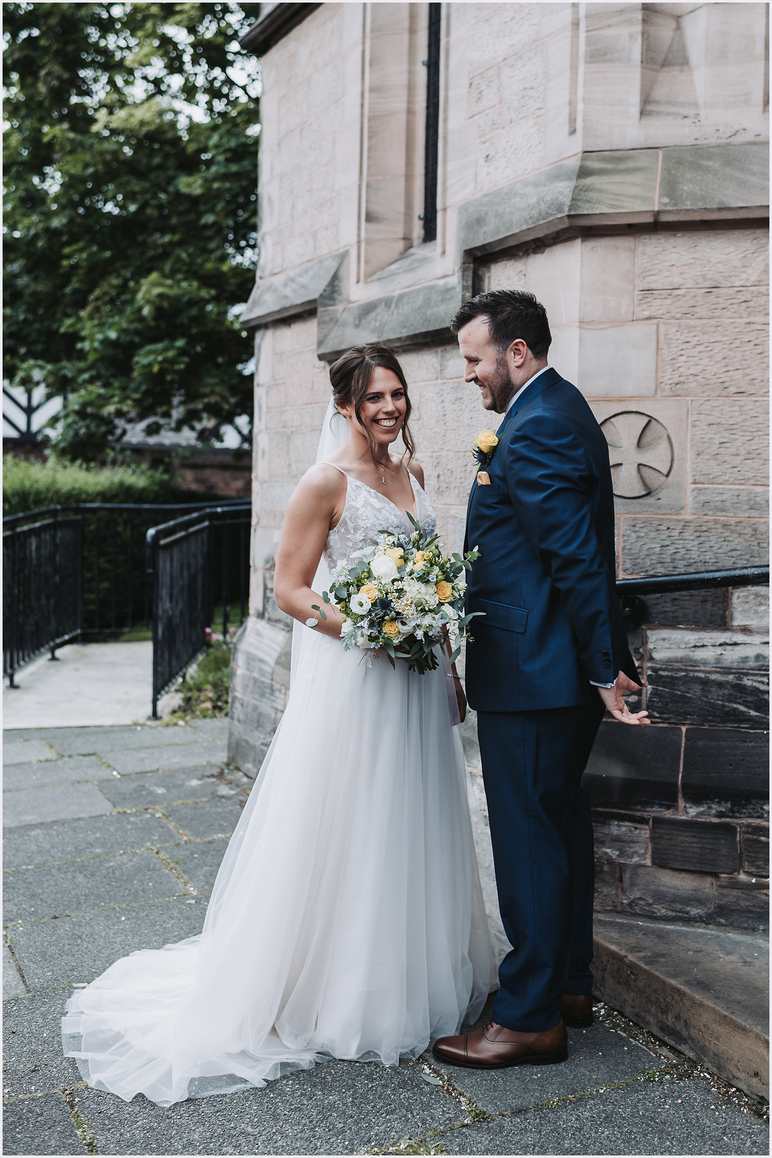 A bride smiles at the camera while her new husband smiles at her outside the church where they have just got married.  Image captured by Helena Jayne Photography.  A wedding photographer covering Cheshire and north Wales