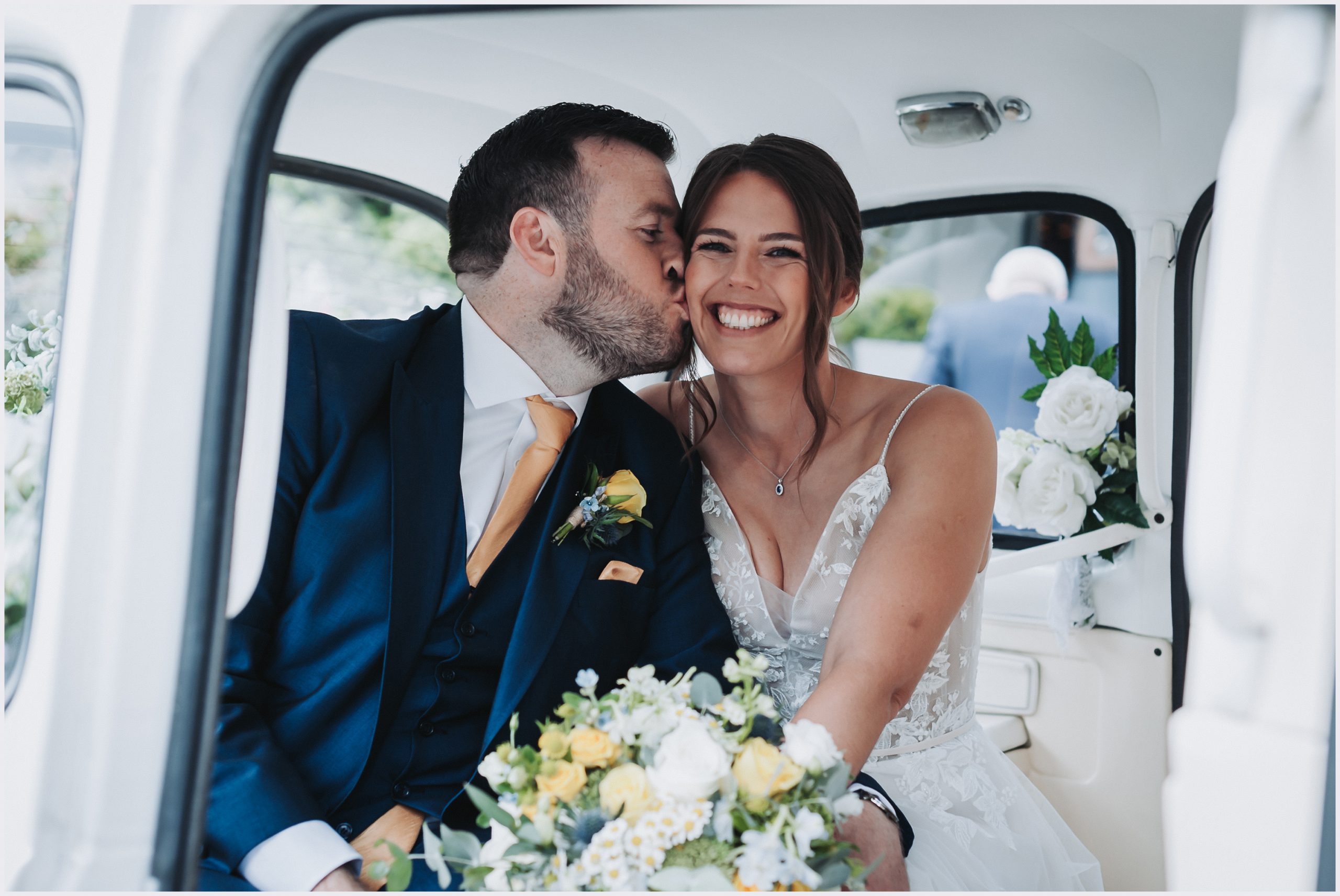 A groom kisses his wife's cheek as she beams at the camera while sitting in their wedding car after thier wedding ceremony.  Image taken by Helena Jayne Photography a wedding photographer based and covering north Wales