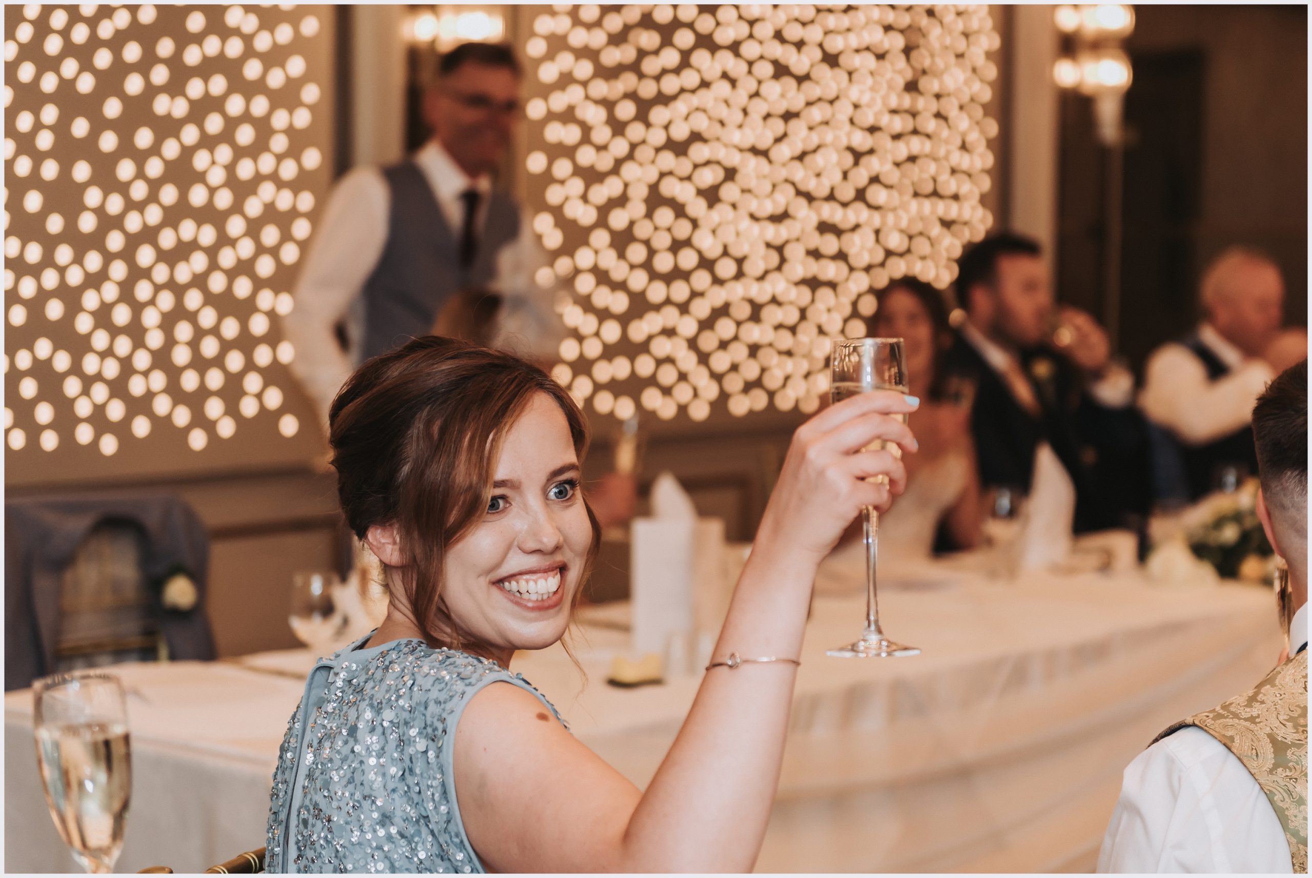 A smiling bridesmaids holds her champagne flute up in the air while making a toast to the happy couple.  Image captured by Helena Jayne Photography a wedding photographer based in Chester
