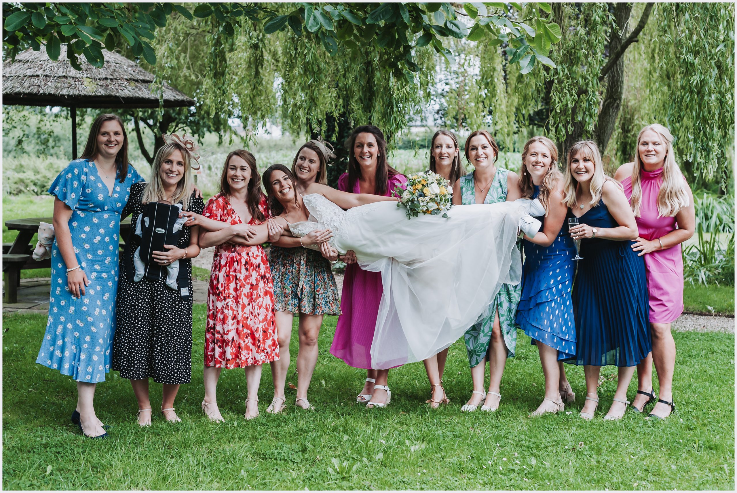 A bride is carried by all of her best friends wearing colourful beautiful dresses in the gorgeous gardens at The Grosvenor Pulford Hotel and Spa.  Image captured by Helena Jayne Photography