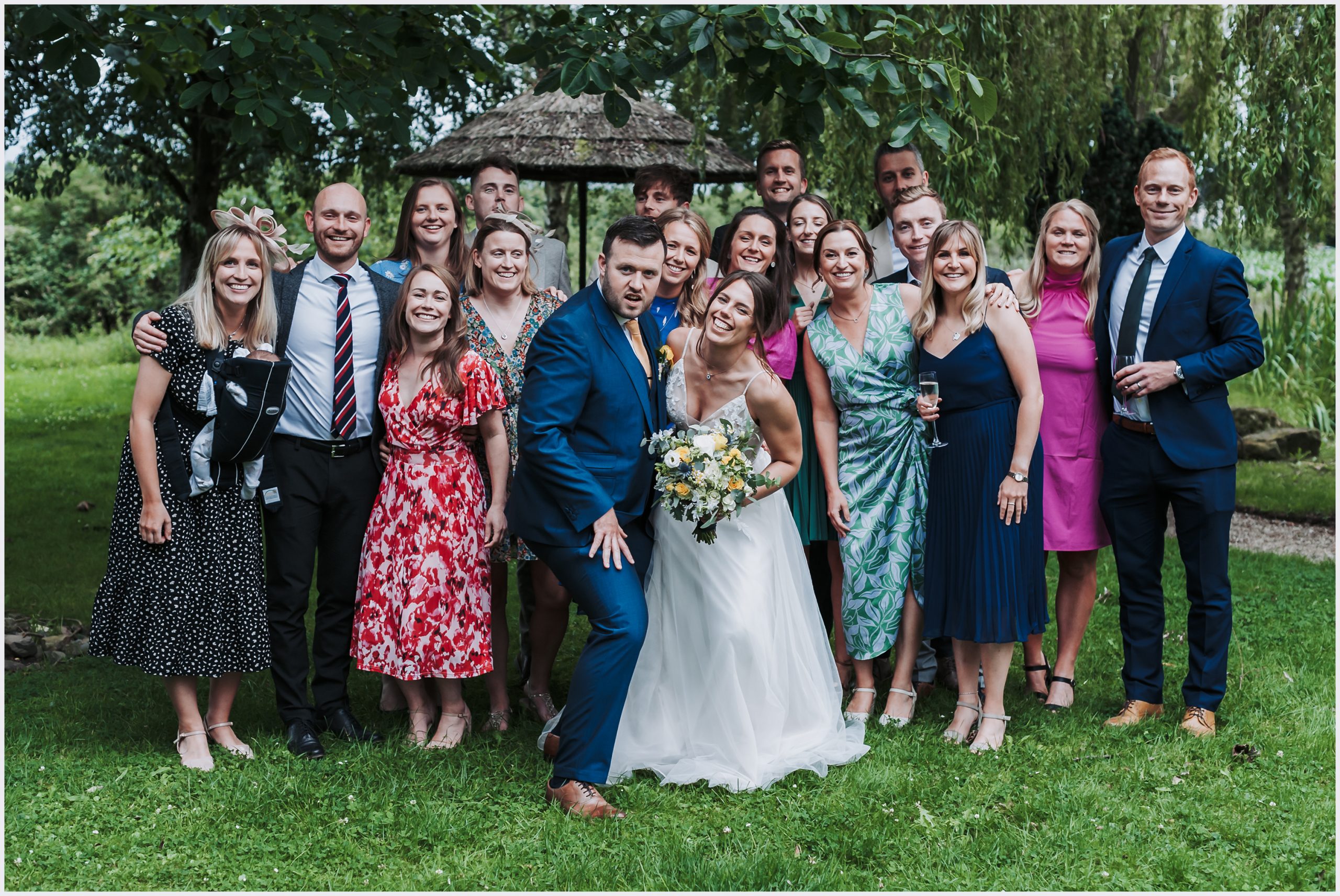 A groom pulls a funny face with his smiling bride during a group shot of all of their friends.  Image captured by Chester wedding photographer Helena Jayne Photography
