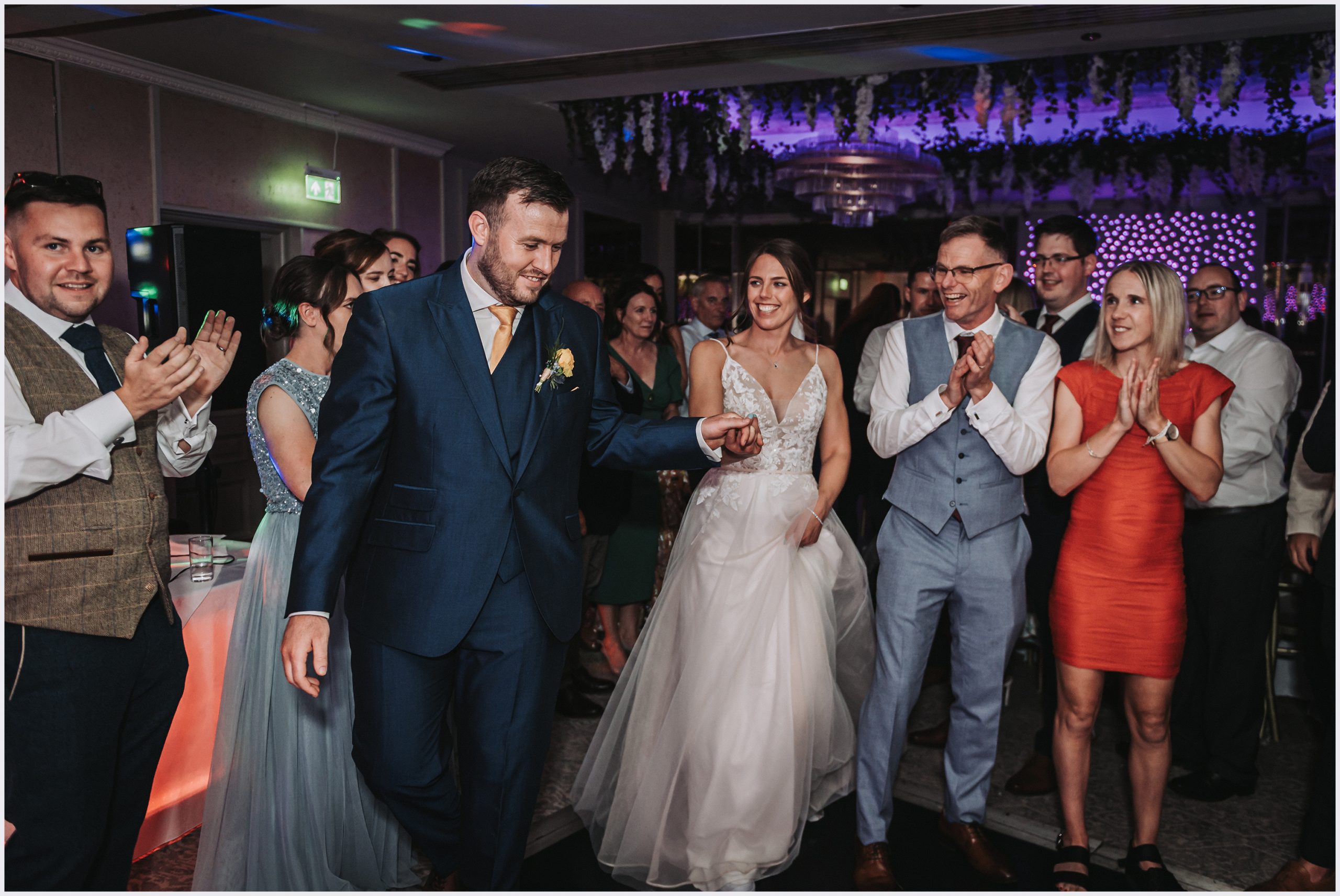 A proud groom leads his beautiful bride on to the dance floor to dance their first dance as man wife as their guests applaud and cheer.  Image captured by north wales based wedding photographer Helena Jayne Photography
