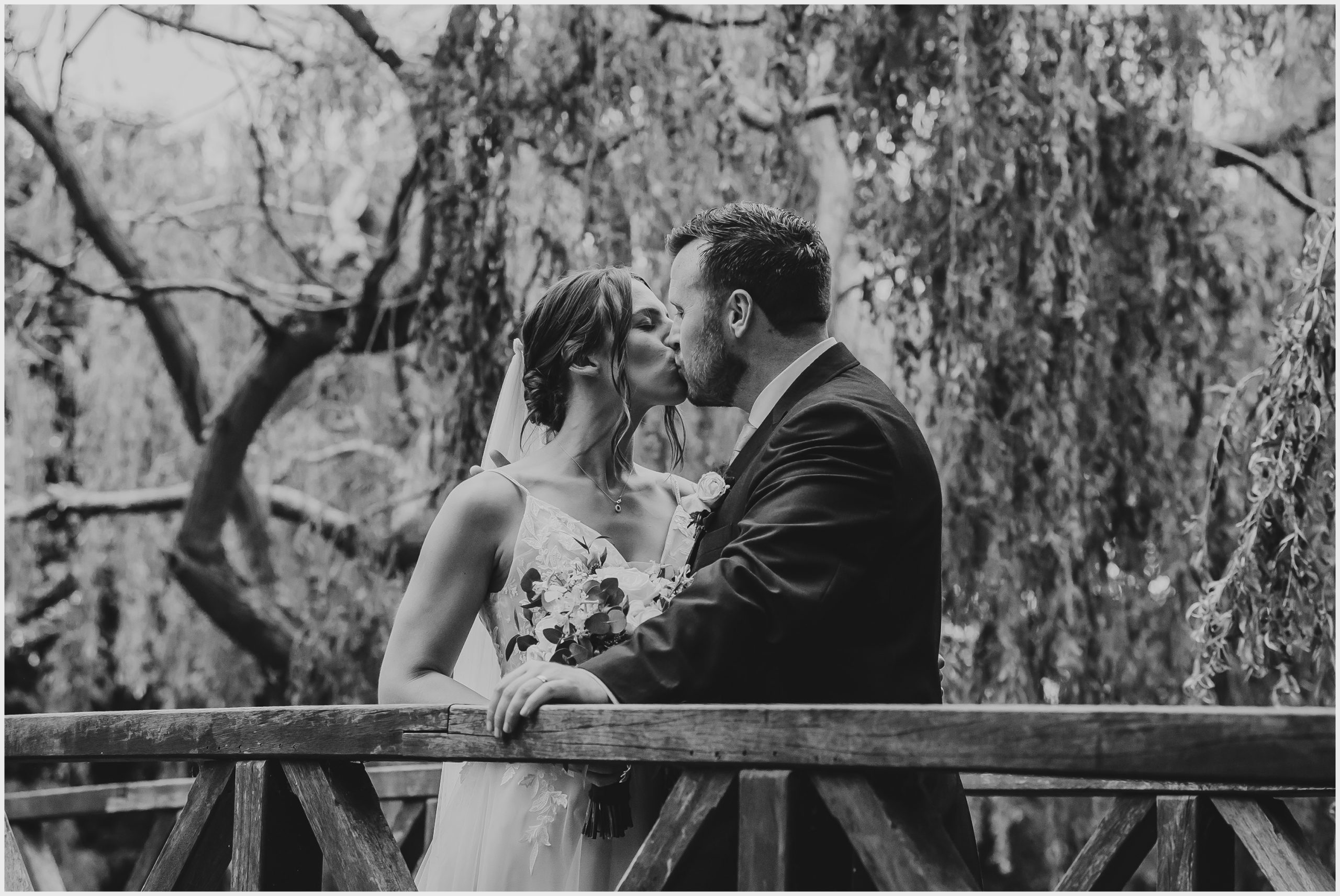 A beautiful bride and groom kiss on a bridge with lush willow trees in the background.  Image captured by Helena Jayne Photography a wedding photographer based in north Wales