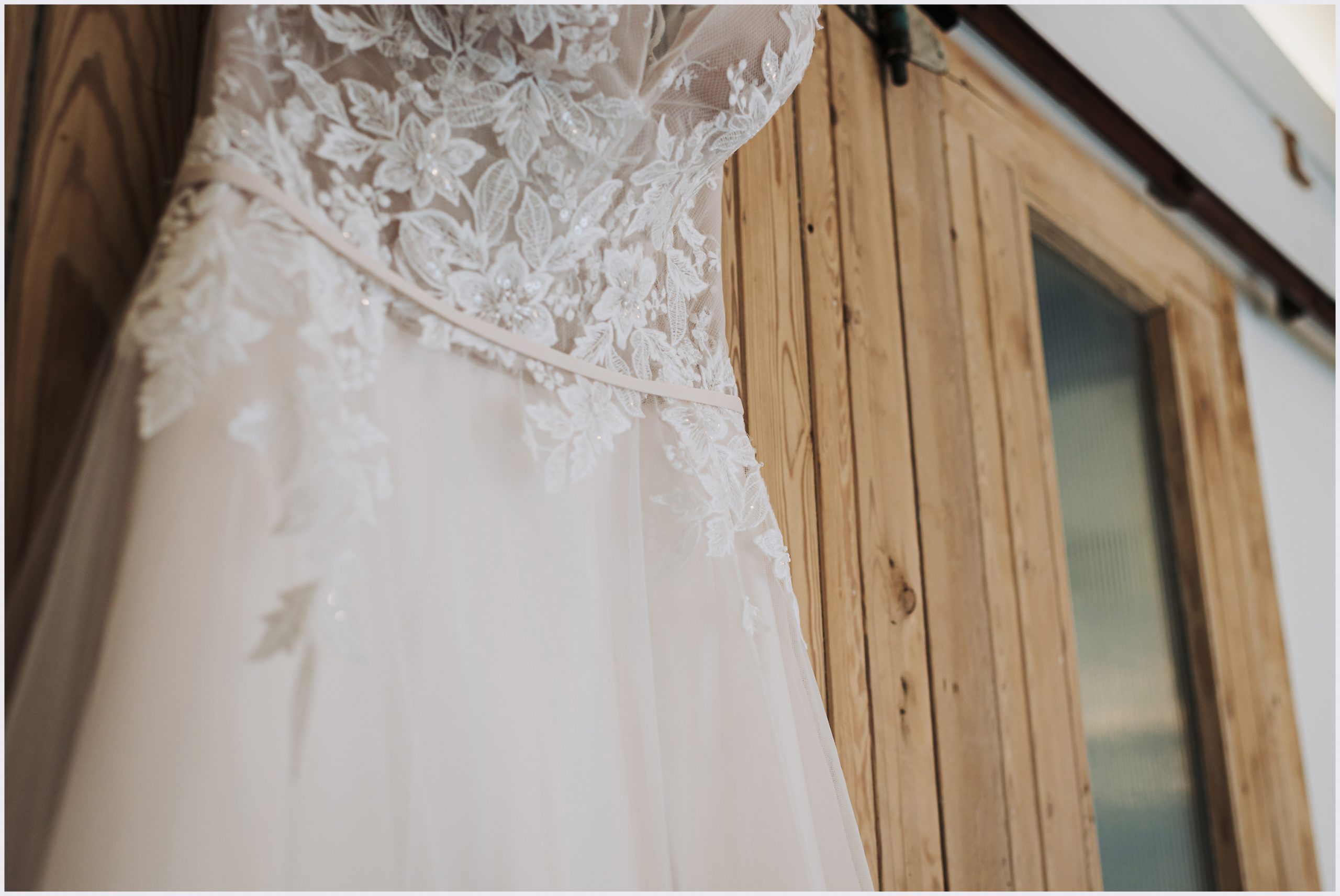 A bride's wedding gown hangs against rustic barn doors showing the lace detail.