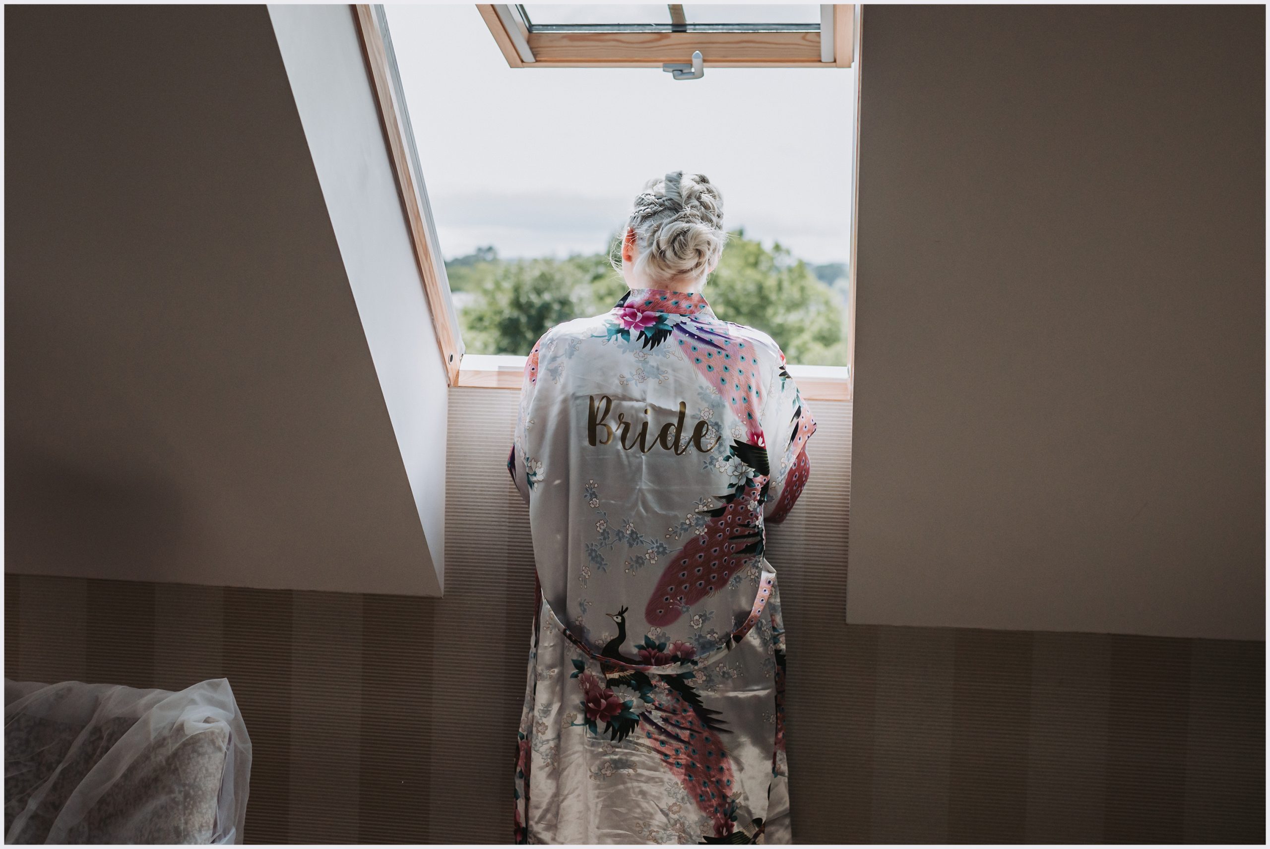 A bride looking out of the window wearing her bridal dressing gown on the morning of her wedding.  Image captured by Helena Jayne Photography, Cheshire wedding photographer