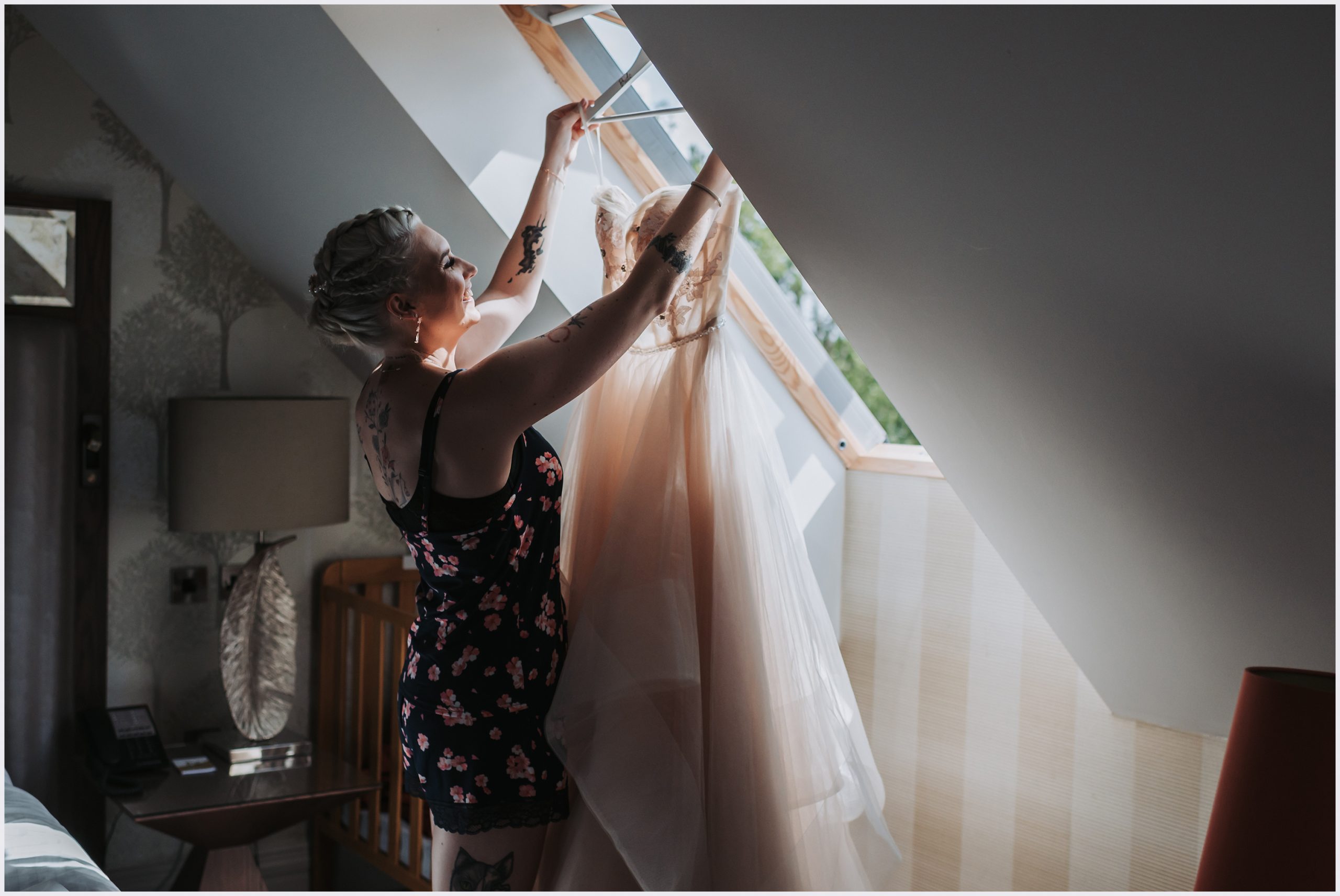 A beautiful bride lifts her wedding dress of the hanger as sun shines through the wondow on the morning of her wedding.  The bride is smiling and full of happiness as she gets ready.  Image captured by Helena Jayne Photography.  A north Wales based wedding photographer