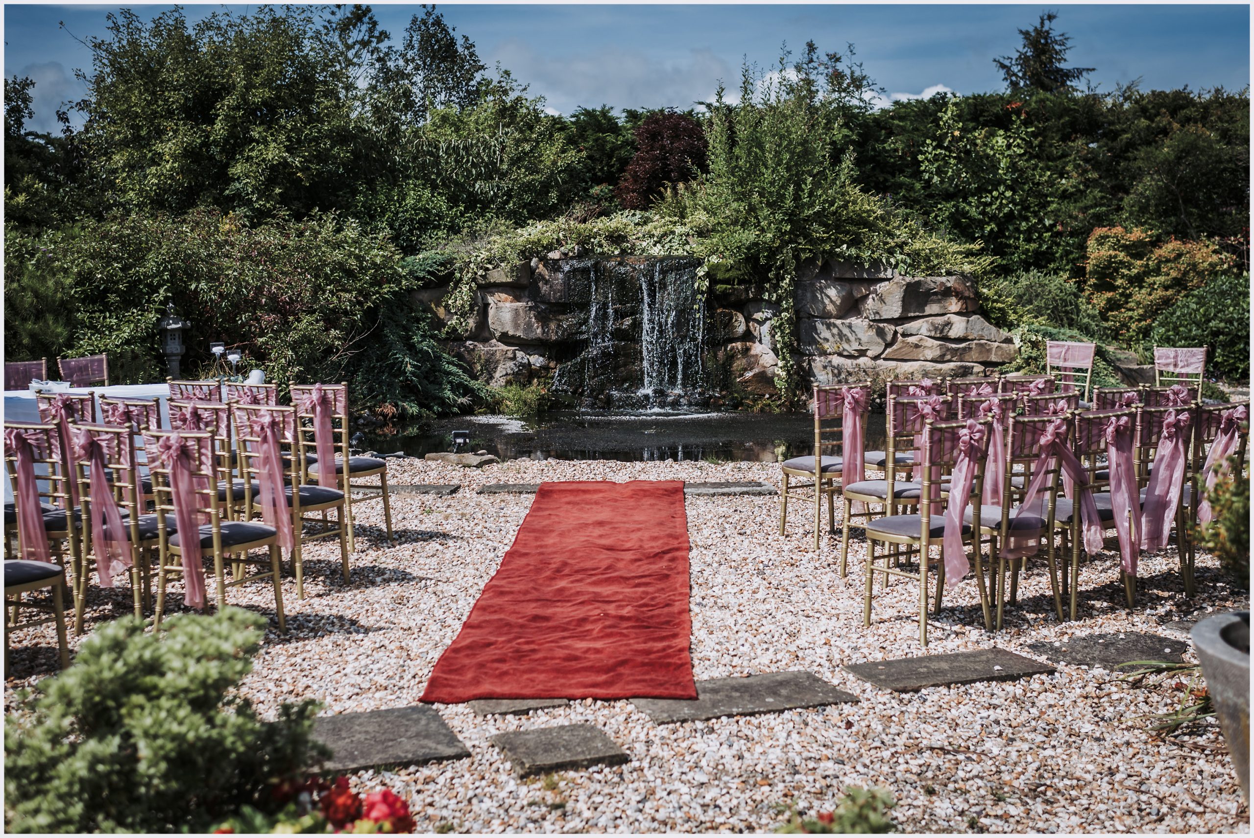 An out door ceremony scene taken at The Grosvenor Pulford Hotel and Spa.  Pale pink sashes are tied in bows on the back of the chairs and a waterfall is in the background.  Image captured by Helena Jayne Photography at the Grosvenor Pulford Hotel and Spa