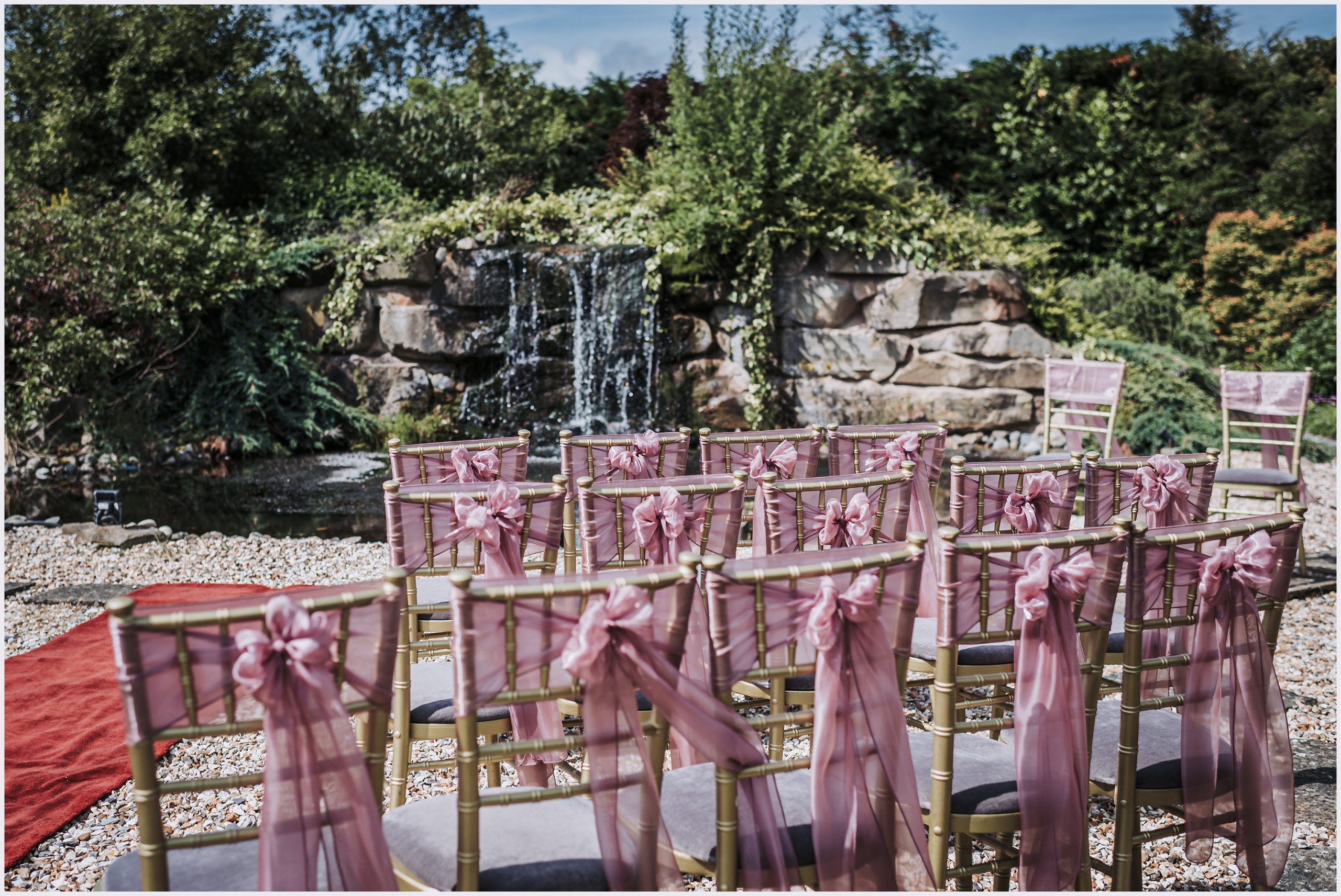 A close up shot of the back of chairs with pale pink sashes tied in bows on the back of them.  The sun is shining and a water feature is in the background.  Image captured at the Grosvenor Pulford Hotel and Spa by north Wales based wedding photographer helena Jayne Photography
