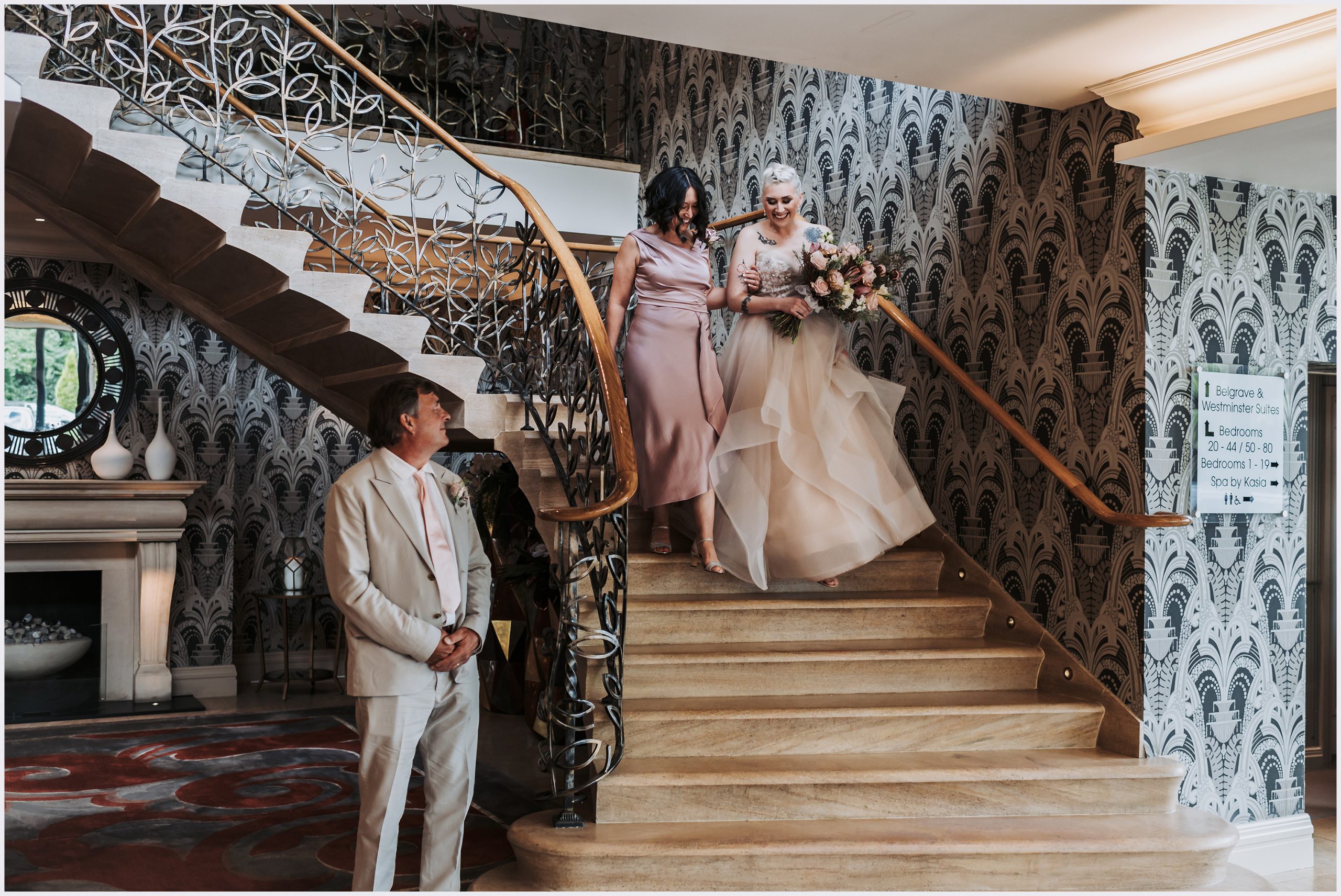 A bride and her mother walk down a the gorgeous staircase at the Grosvenor Pulford Hotel and Spa.  The bride's father waits for them at the bottom of the stira.  Image captured by Helena Jayne Photography, Cheshire based wedding photographer