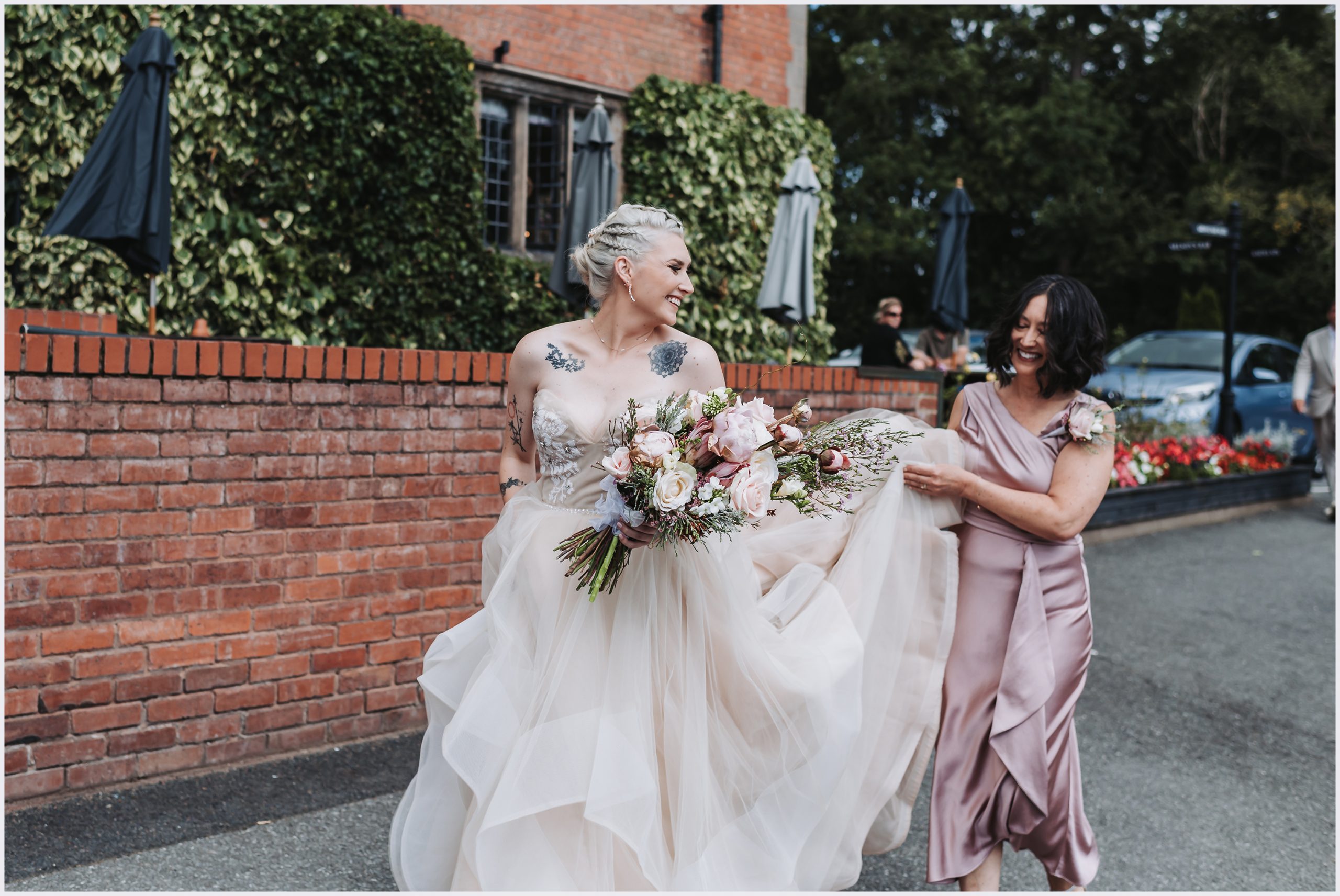 A bride and her mother walking towards the wedding ceremony.  The bride looks back at her mum smiling while her mum holds up the wedding dress.  Image captured by Helena Jayne Photography.  North Wales based wedding photographer