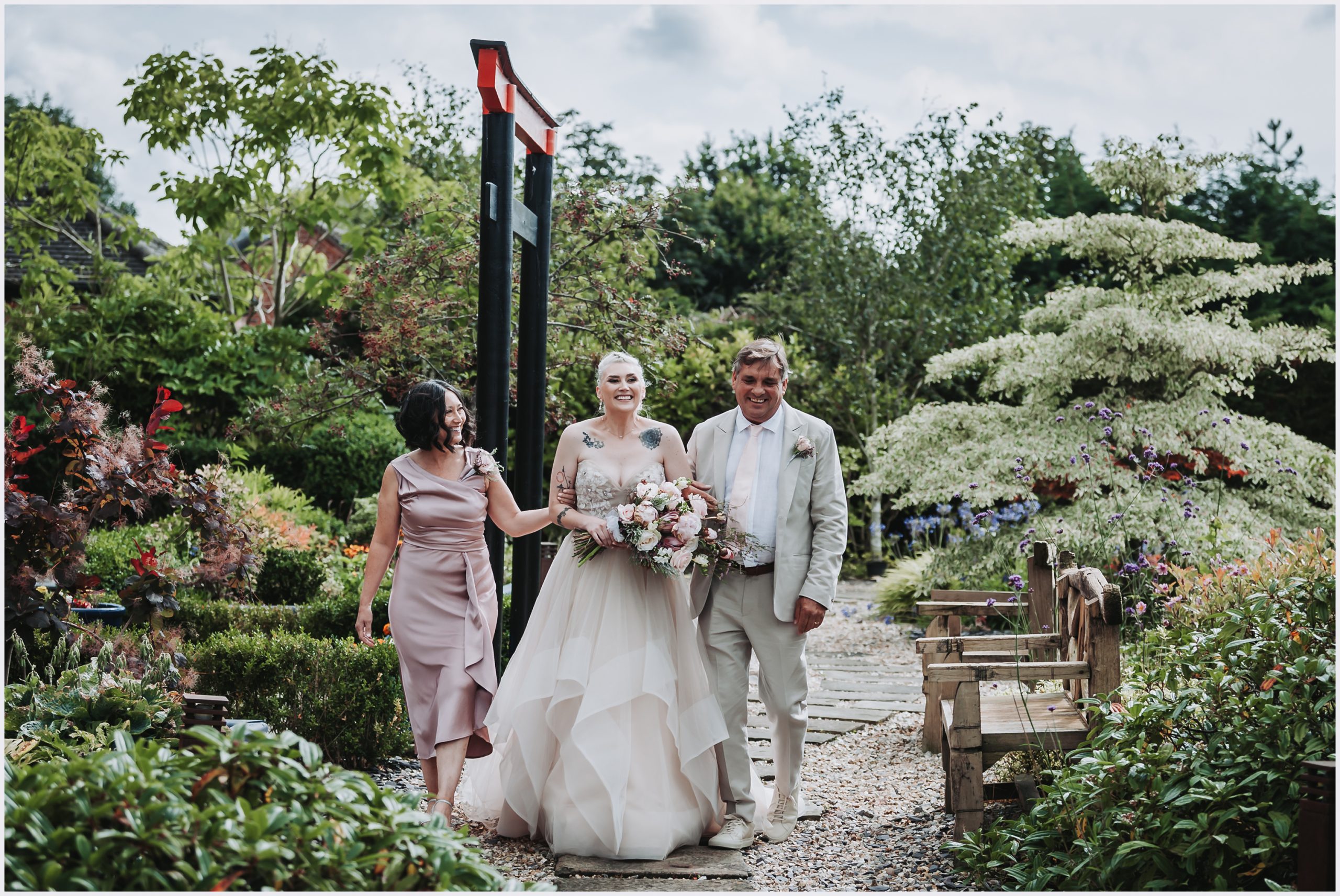 A beaming beautiful bride walks with her parents by her side.  The bride is carrying a beautiful bouquet of flowers and all three are smiling widely as they walk towards the outdoor wedding ceremony at The Grosveor Pulford Hotel and Spa.  Image captured by Helena Jayne Photography a north Wales based wedding photographer