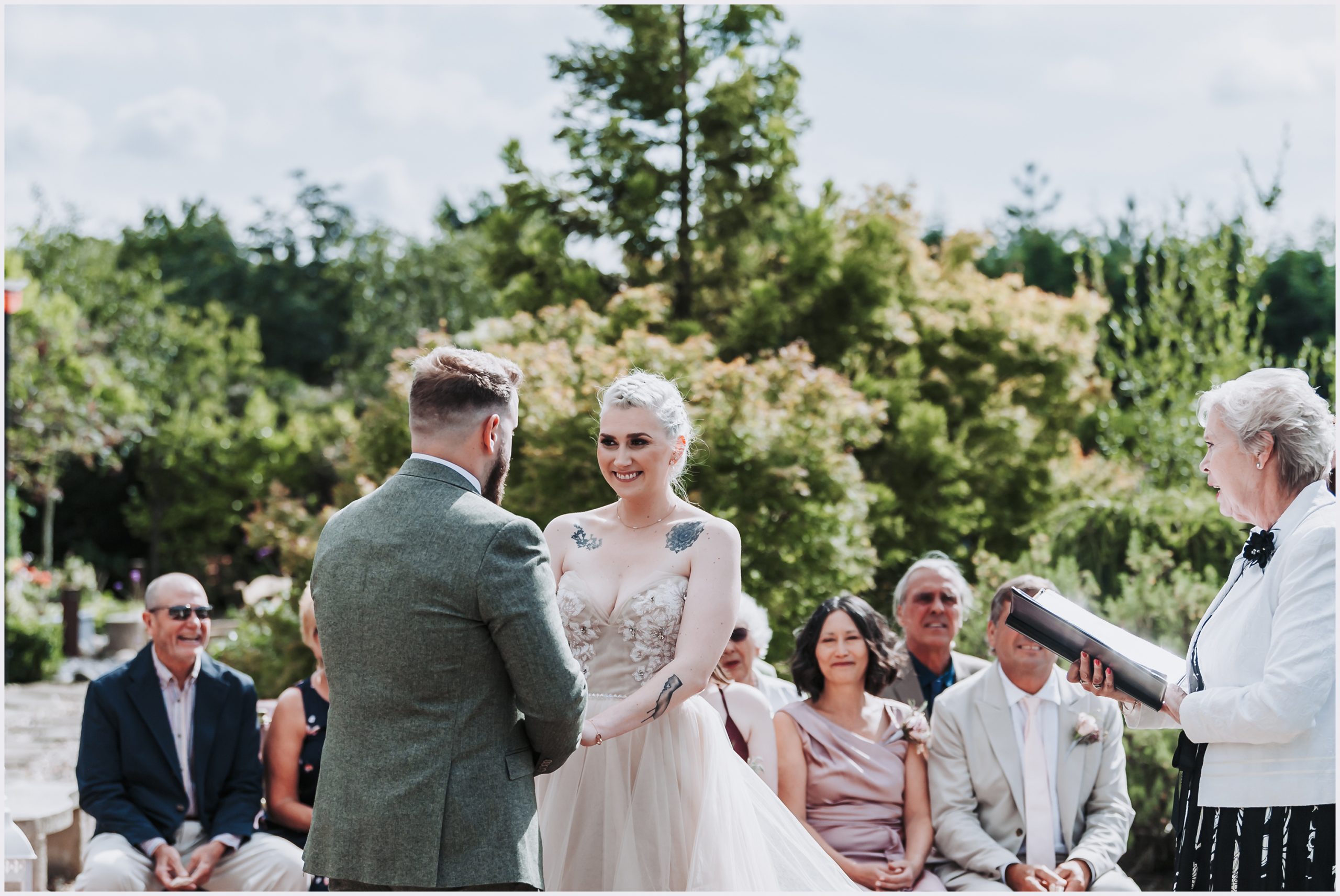 A bride smiles coyly at her fiance during their wedding ceremony.  The registrar who is marrying them stands on the right and guests are smiling happily as the vows are said during an outdoor ceremony at The Grosvenor Pulford Hotel and Spa