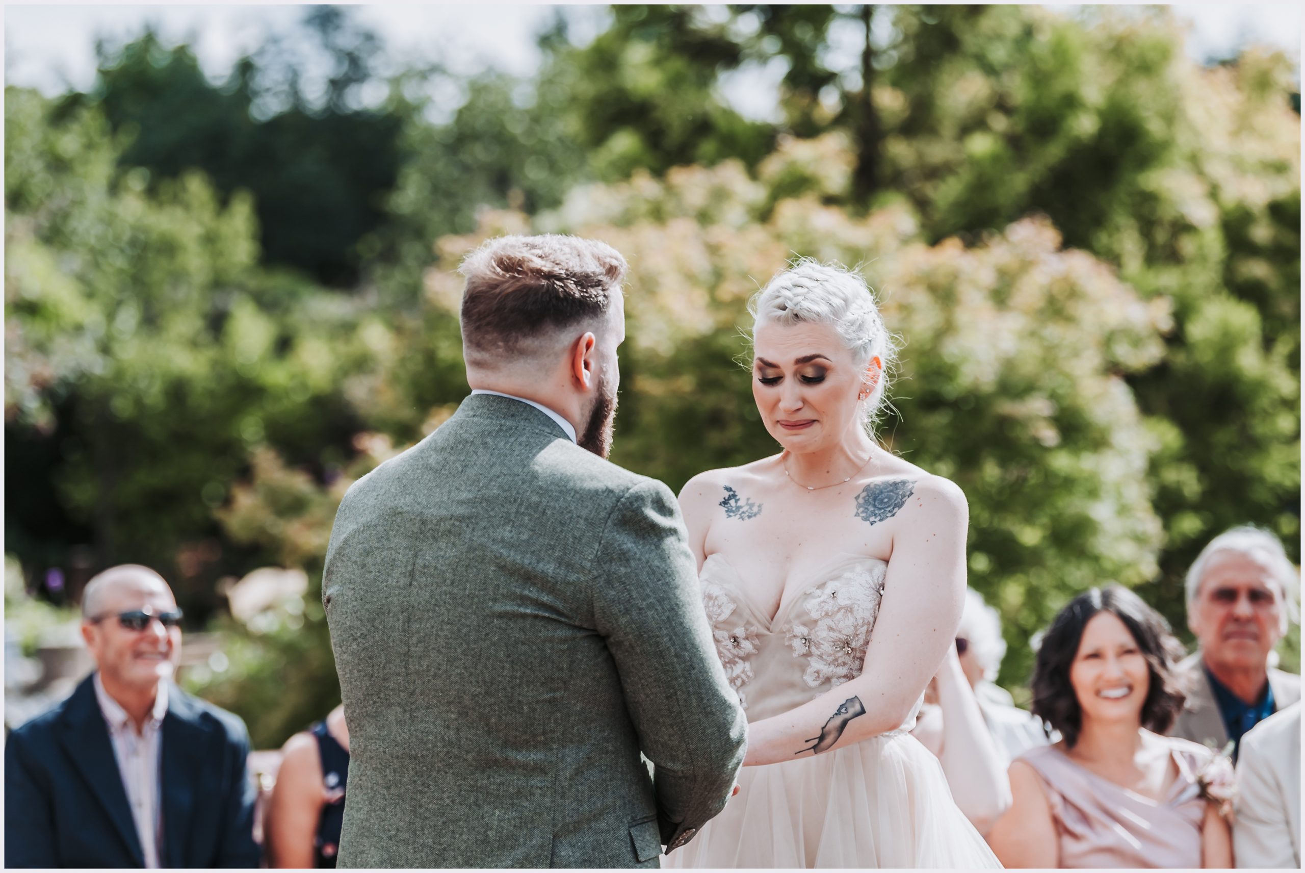 An emotional bride holding back tears of happiness as her fiance repeats his vows during their wedding ceremony.  Guests are in the background smiling happily as they witness the ceremony.  Image captured by Helena Jayne Photography, Cheshire wedding photographer