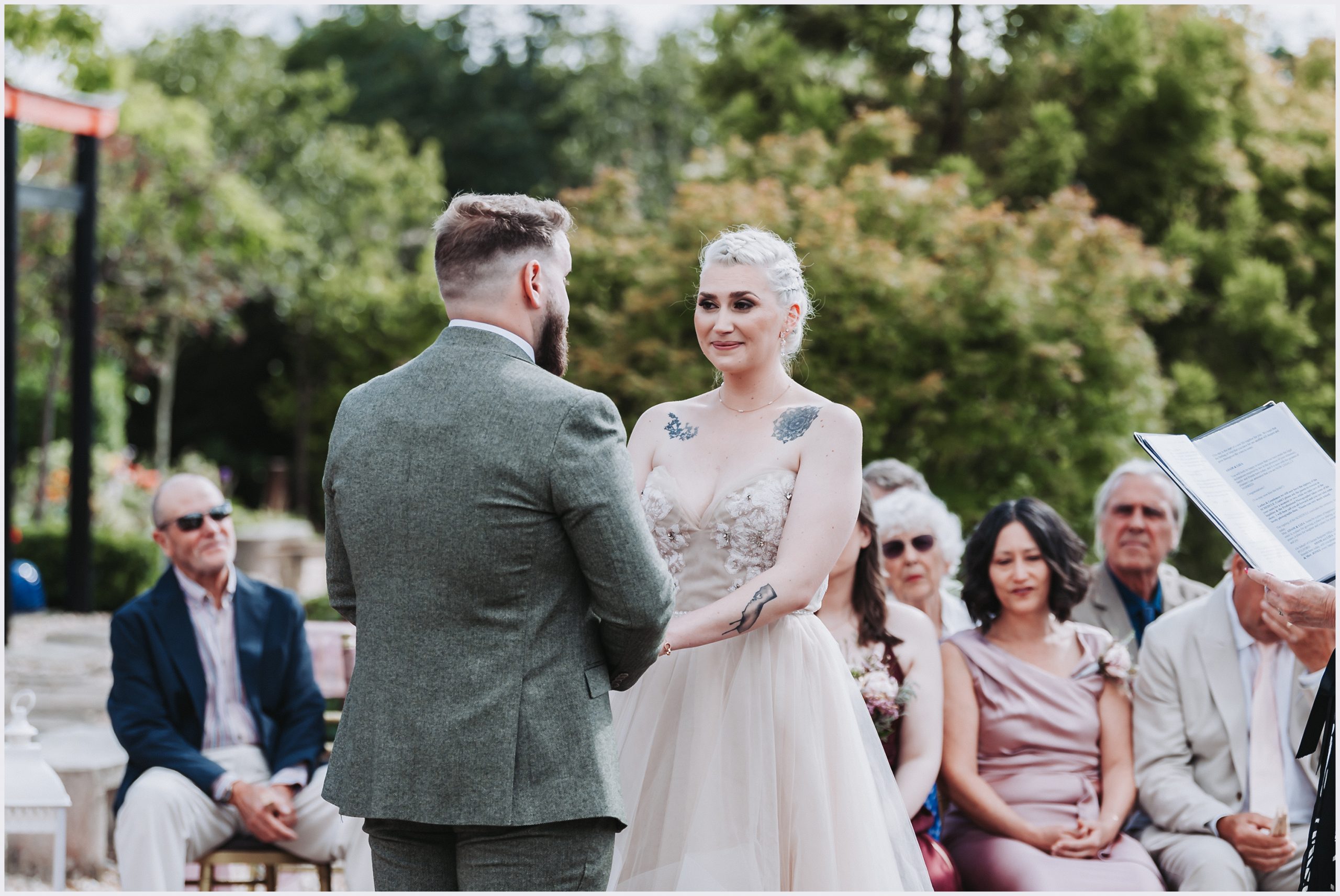 An emotional bride looks intently at her fiance during their outdoor wedding ceremony at The Grosvenor Pulford Hotel and Spa.  Guests are in the background smiling as they listen to the vows.  Image captured by Helena Jayne Photography.