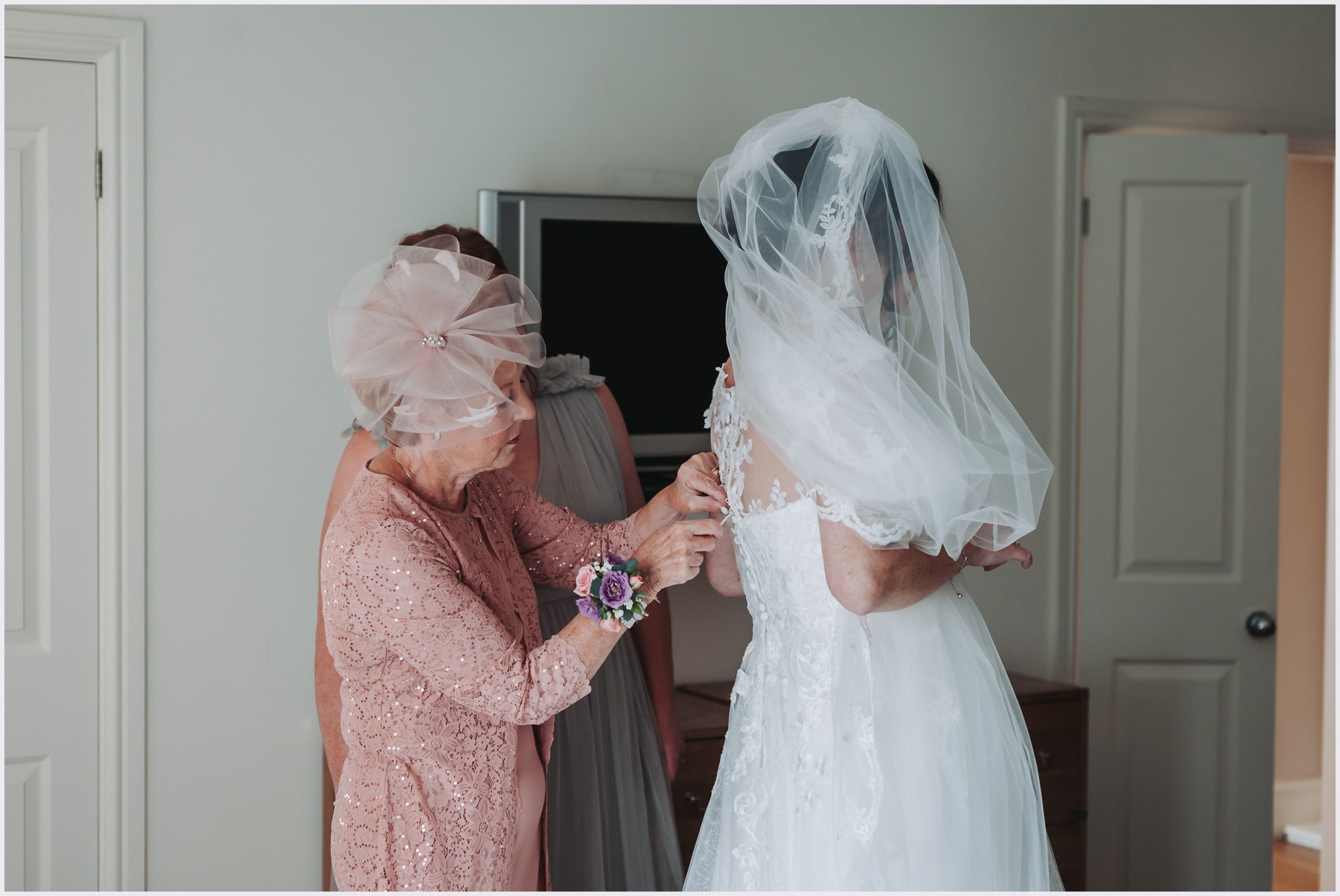 A bride's mother fastens the buttons with a crochet stick on her daughter's wedding dress on the morning of her wedding.  Image captured at Rowton Hall Hotel and Spa in Chester by Cheshire based wedding photographer Helena Jayne Photography