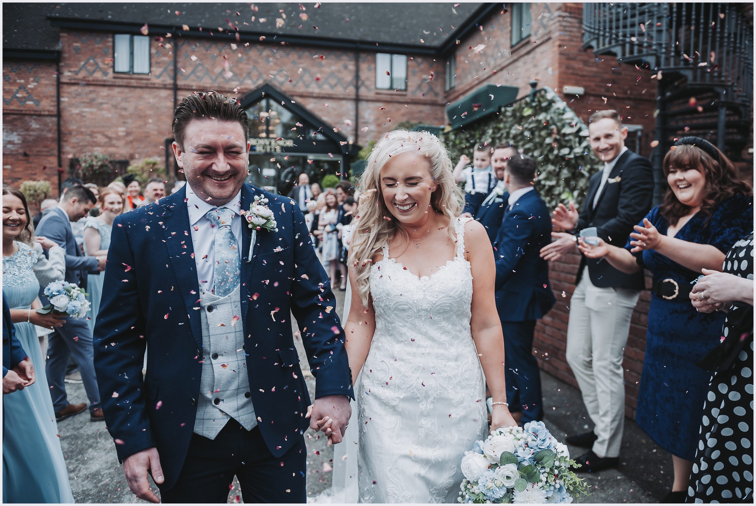 A newly married bride and groom smile and laugh as their guests shower them with confetti outside The Grosvenor Pulford Hotel and Spa.  Image captured by north Wlaes wedding photographer Helena Jayne Photography.