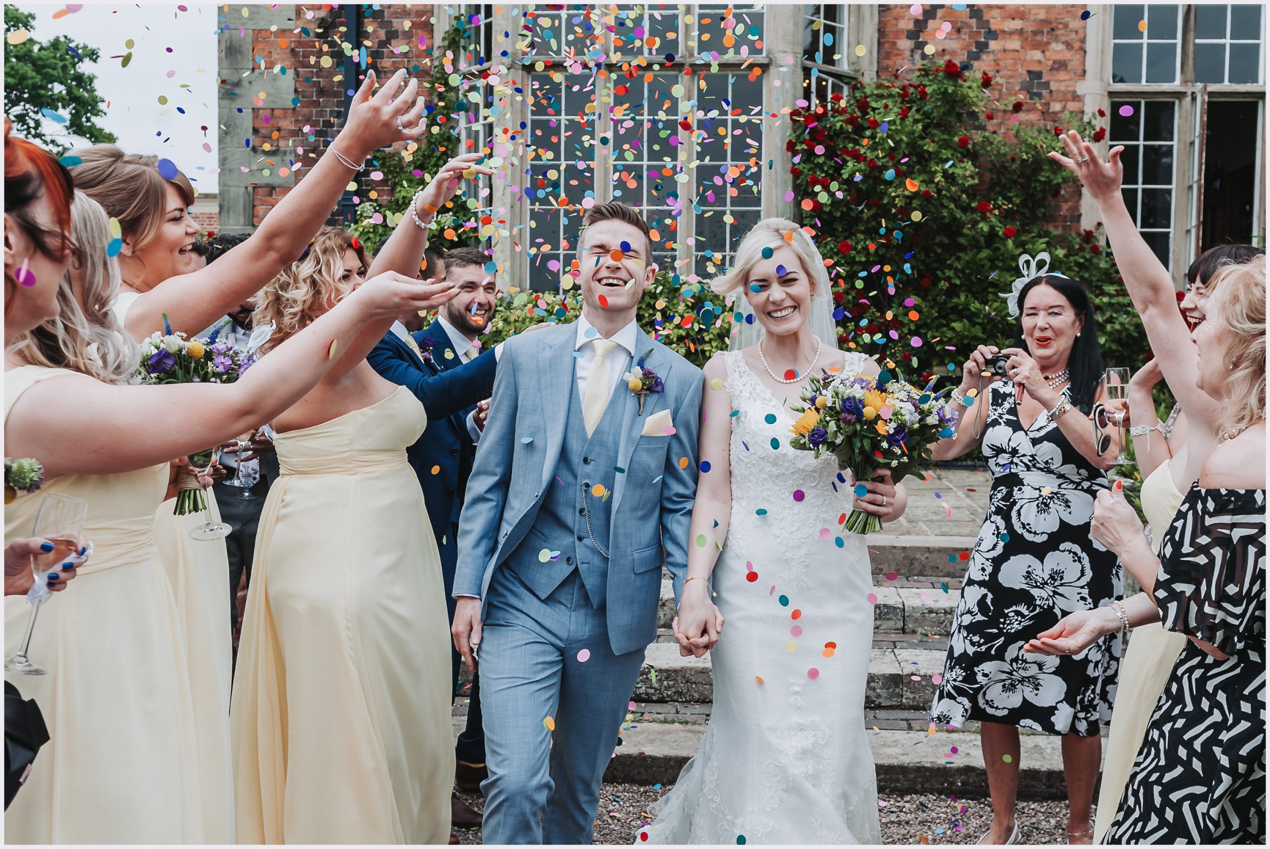 A gorgeous bride and groom smile outside Willington Hall Hotel in Cheshire after getting married.  Guests are throwing brightly colour confetti at them as they walk past.  Image captured by Cheshire Wedding Photographer Helena Jayne Photography.