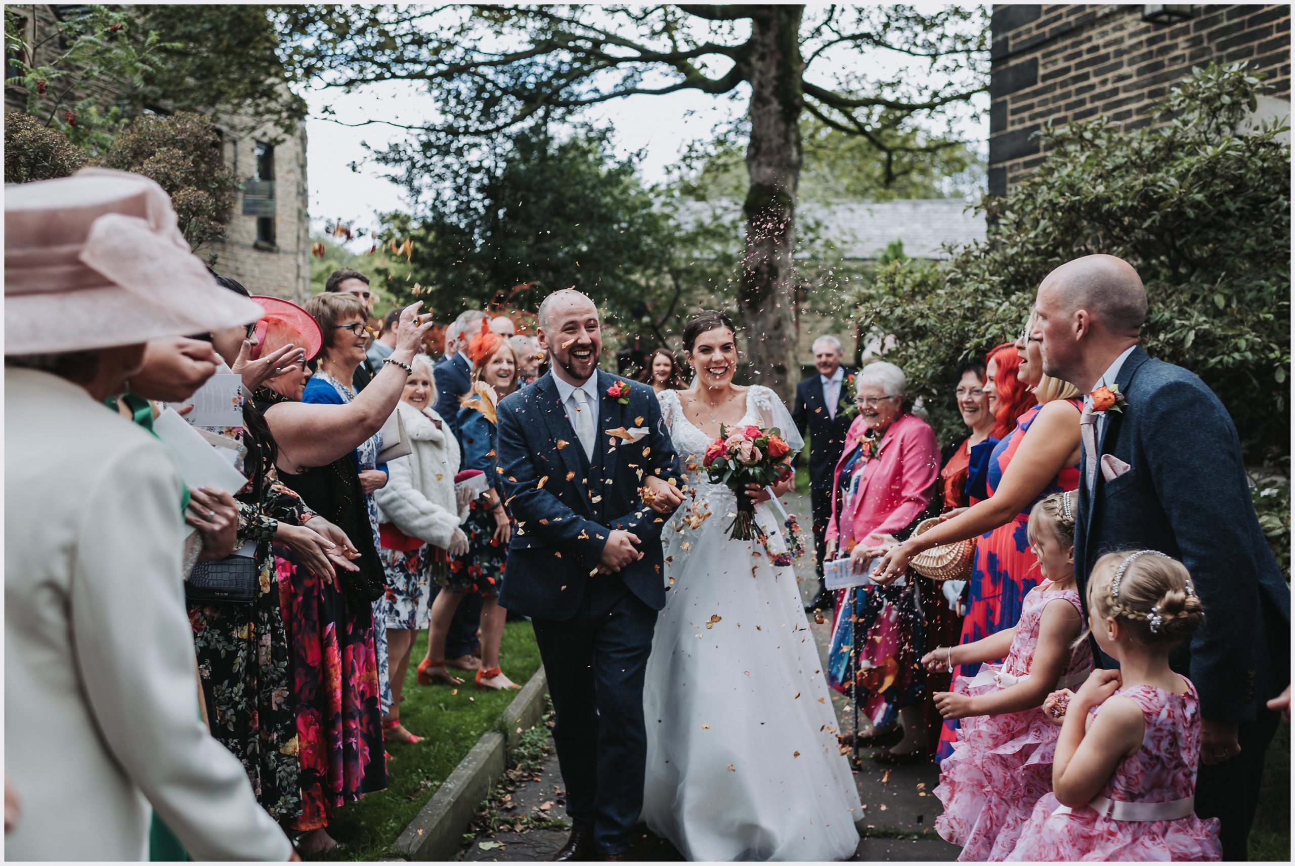 A smiling and laughing newly married couple smile as they walk along the path outside the church where they've just got married.  Their guests are throwing confetti at them as they walk by.  Image captured by Cheshire Wedding photographer Helena Jayne Photography.
